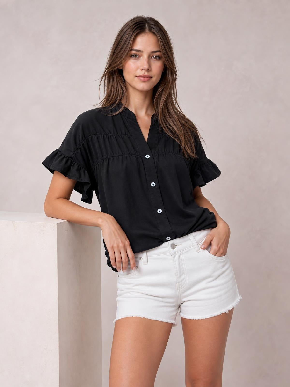 A model poses with one hand in her shorts pocket, wearing a black button-down shirt with a relaxed silhouette, short ruffled sleeves, and a front button placket.
