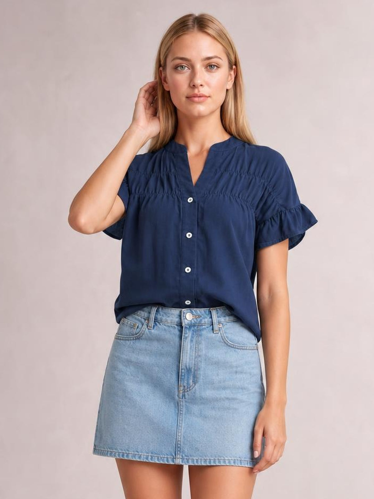 A model poses with one hand near her hair wearing a navy button-down shirt with ruched detailing, ruffled short sleeves, and a relaxed silhouette.