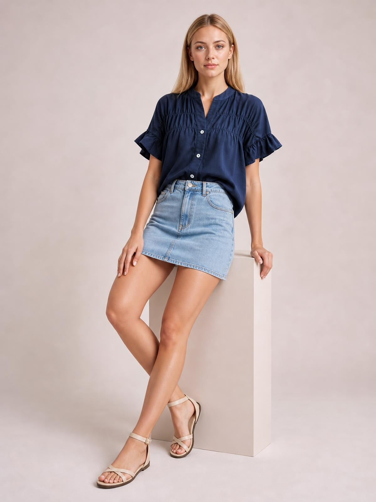Model poses in a navy button-down shirt with short ruffled sleeves, ruched detailing, and a relaxed silhouette styled with a denim skirt.