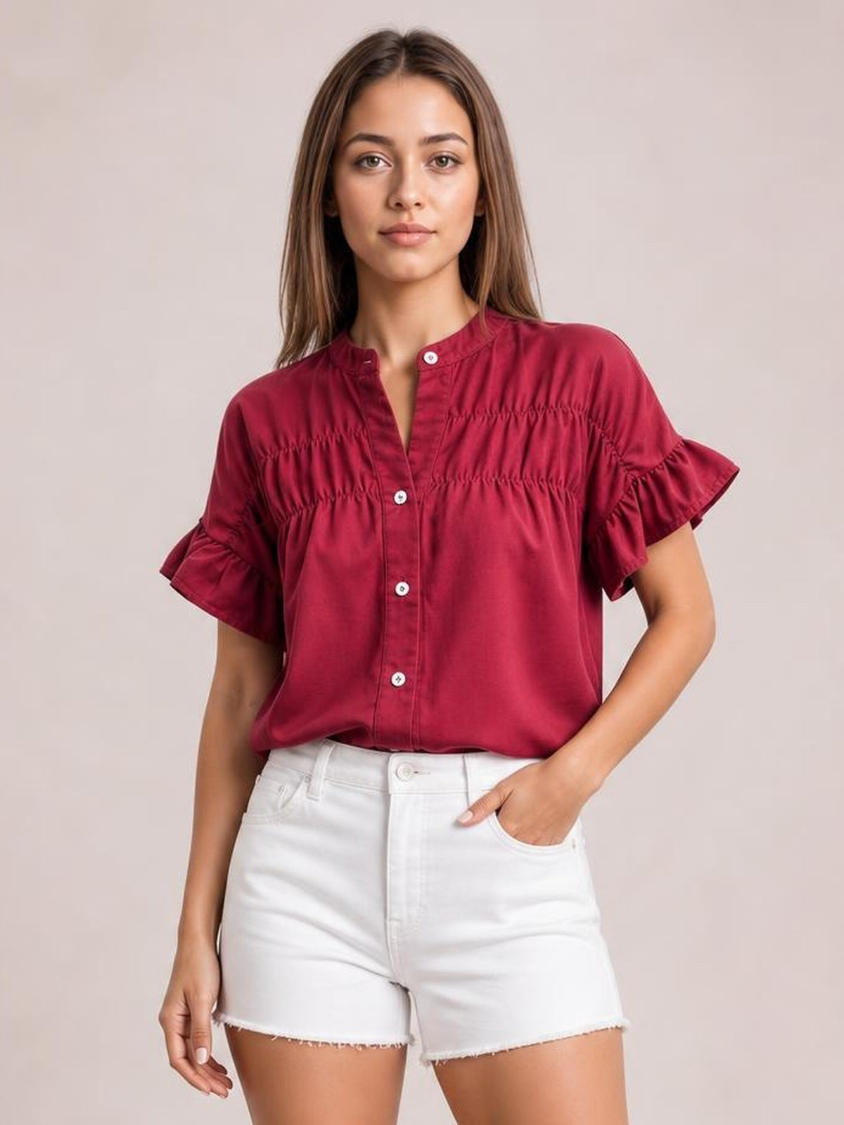 Model wearing a burgundy button-down shirt with short ruffled sleeves, a ruched yoke, and a relaxed silhouette, posing with one hand in her shorts pocket.