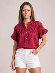 Model wearing a burgundy button-down shirt with short ruffled sleeves, a ruched yoke, and a relaxed silhouette, posing with one hand in her shorts pocket.