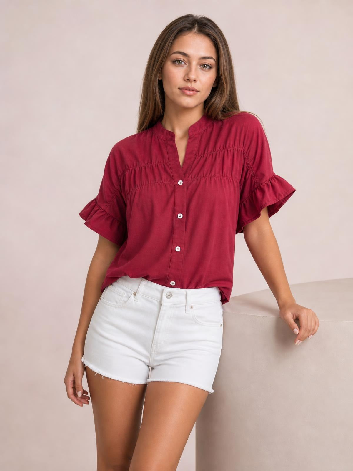A model poses in a burgundy button-down shirt with short ruffled sleeves, ruched seam detailing, a relaxed silhouette, and a curved hem.