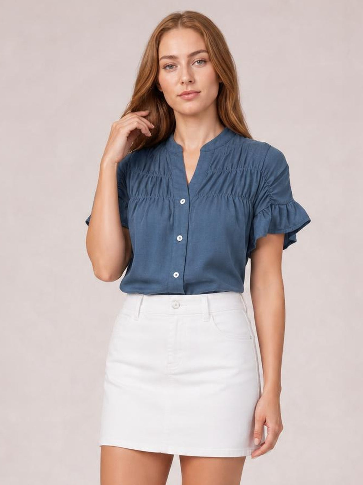 Model wearing a blue button-down short sleeve shirt with ruched detailing, a split neckline, and layered ruffled sleeves, posed with one hand near her hair.