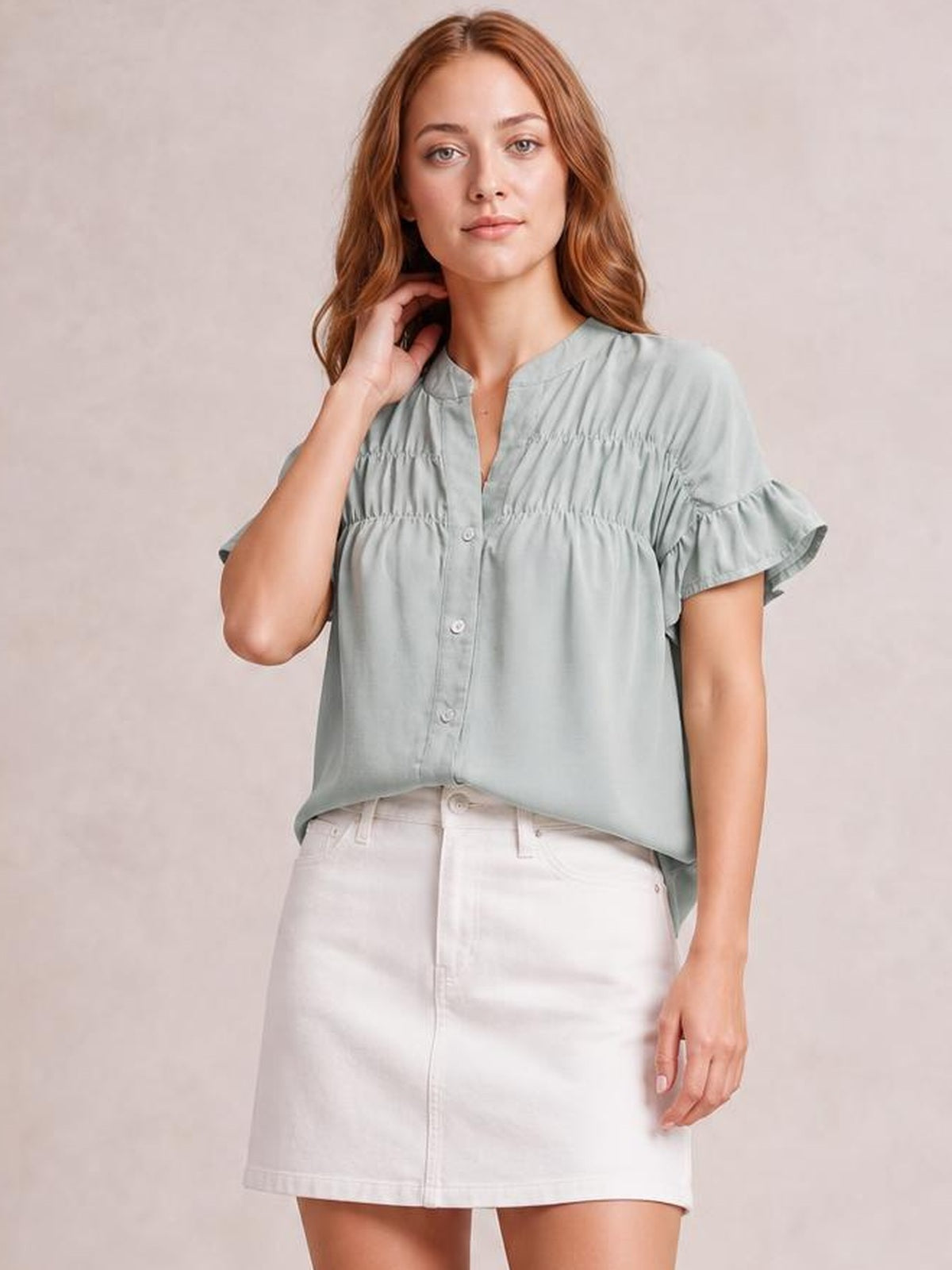 A model poses in a pale sage-green button-down shirt with short ruffled sleeves, ruched horizontal detailing, and a relaxed, loose silhouette.