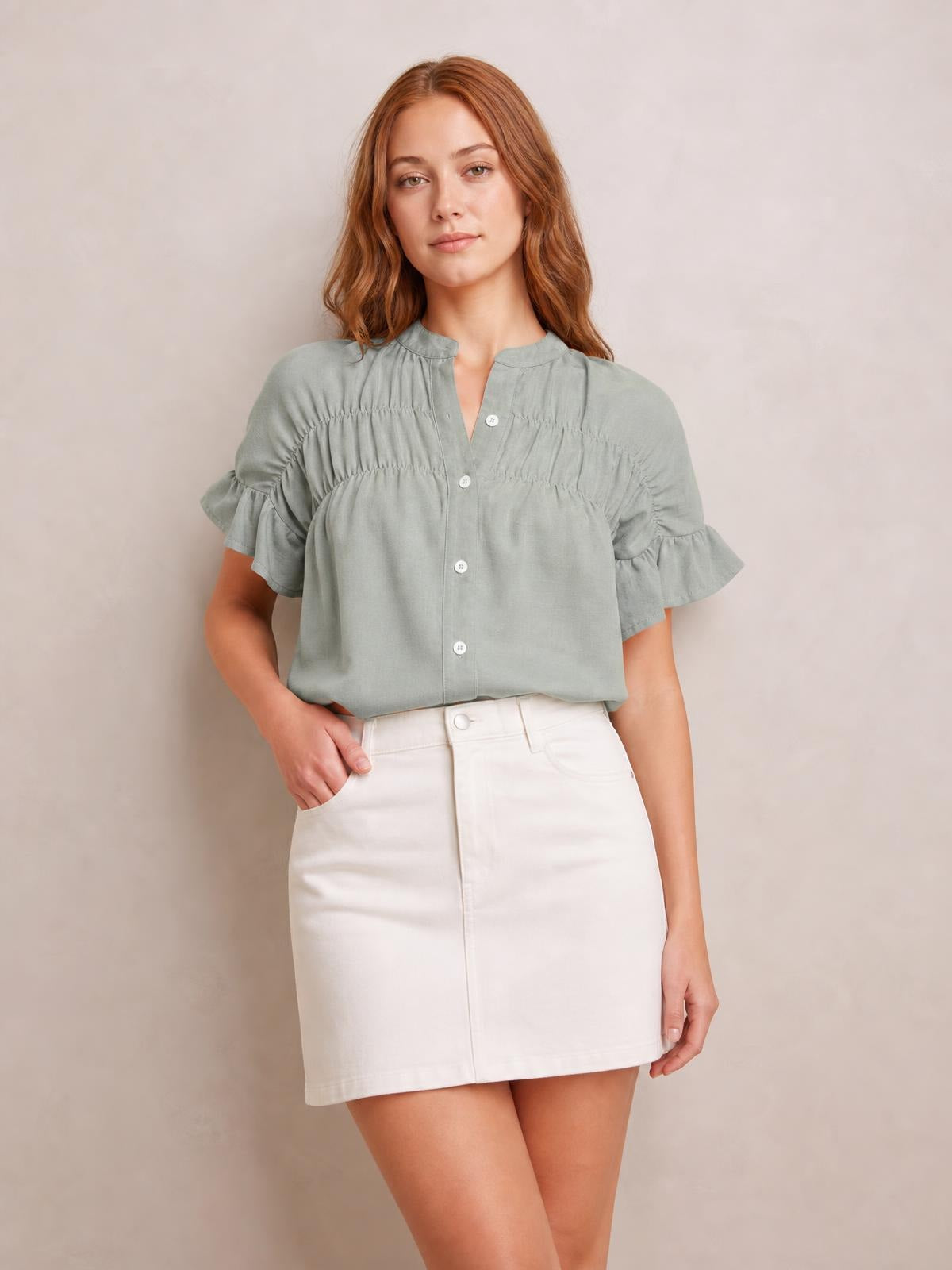 A model wears a sage green button-down shirt with short ruffled sleeves, a ruched yoke, band collar, and relaxed silhouette, posing with one hand in her pocket.