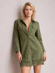 A model wears an olive-green button-down shirt dress with a collared neckline, long sleeves, chest pockets, relaxed silhouette, and lace eyelet trim at the cuffs and hem.