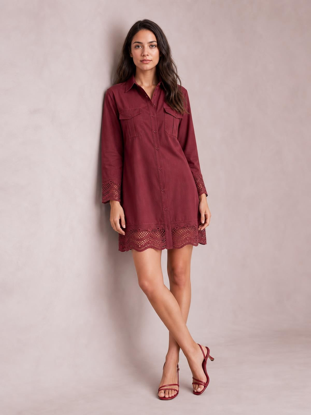 A model poses with crossed legs in a burgundy button-down shirt dress featuring long sleeves, flap chest pockets, and lace eyelet trim at the cuffs and hem.