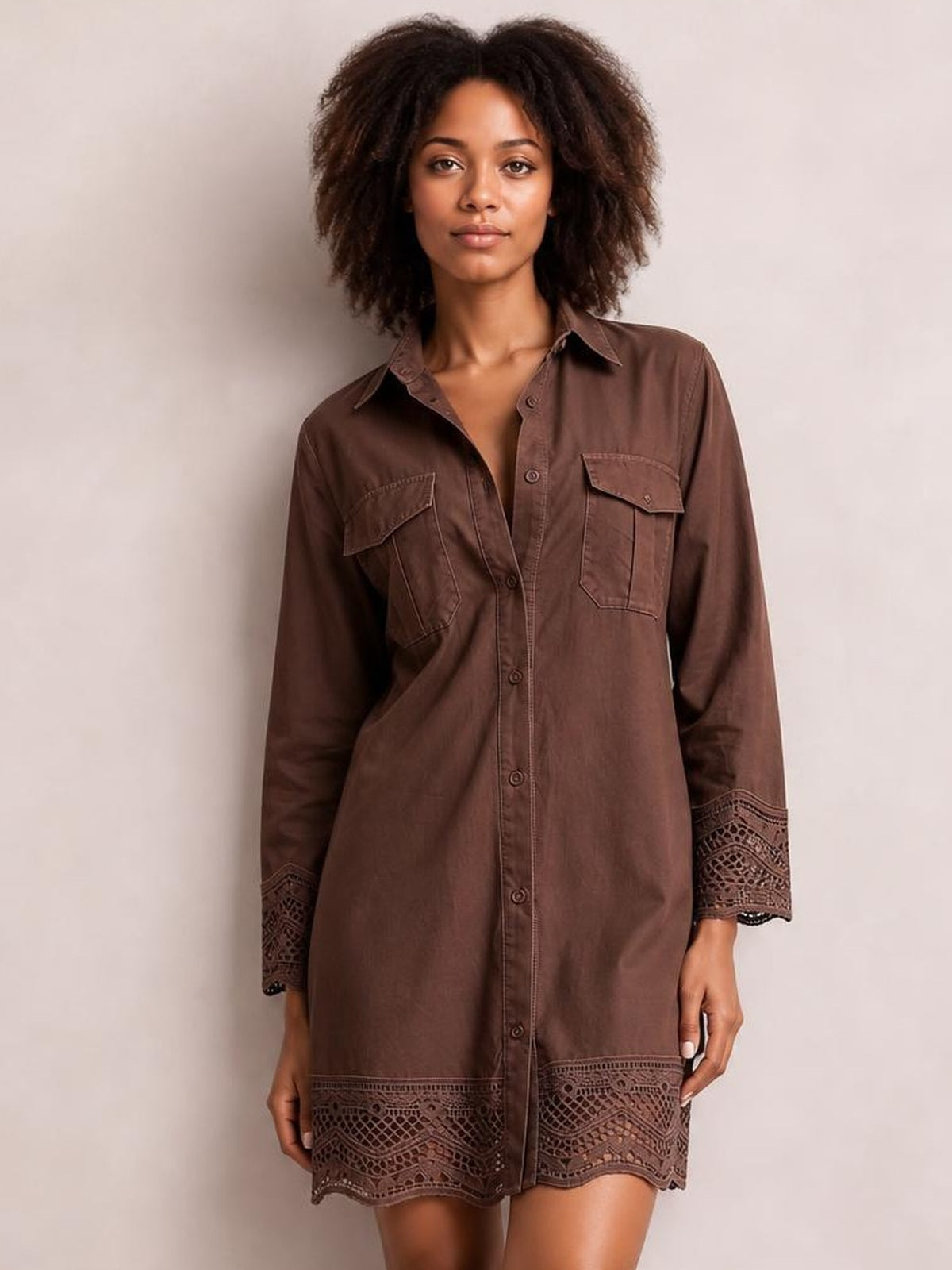 A model stands in a brown button-down shirt dress with a collared neckline, long sleeves, chest pockets, and intricate lace trim at the cuffs and hem.