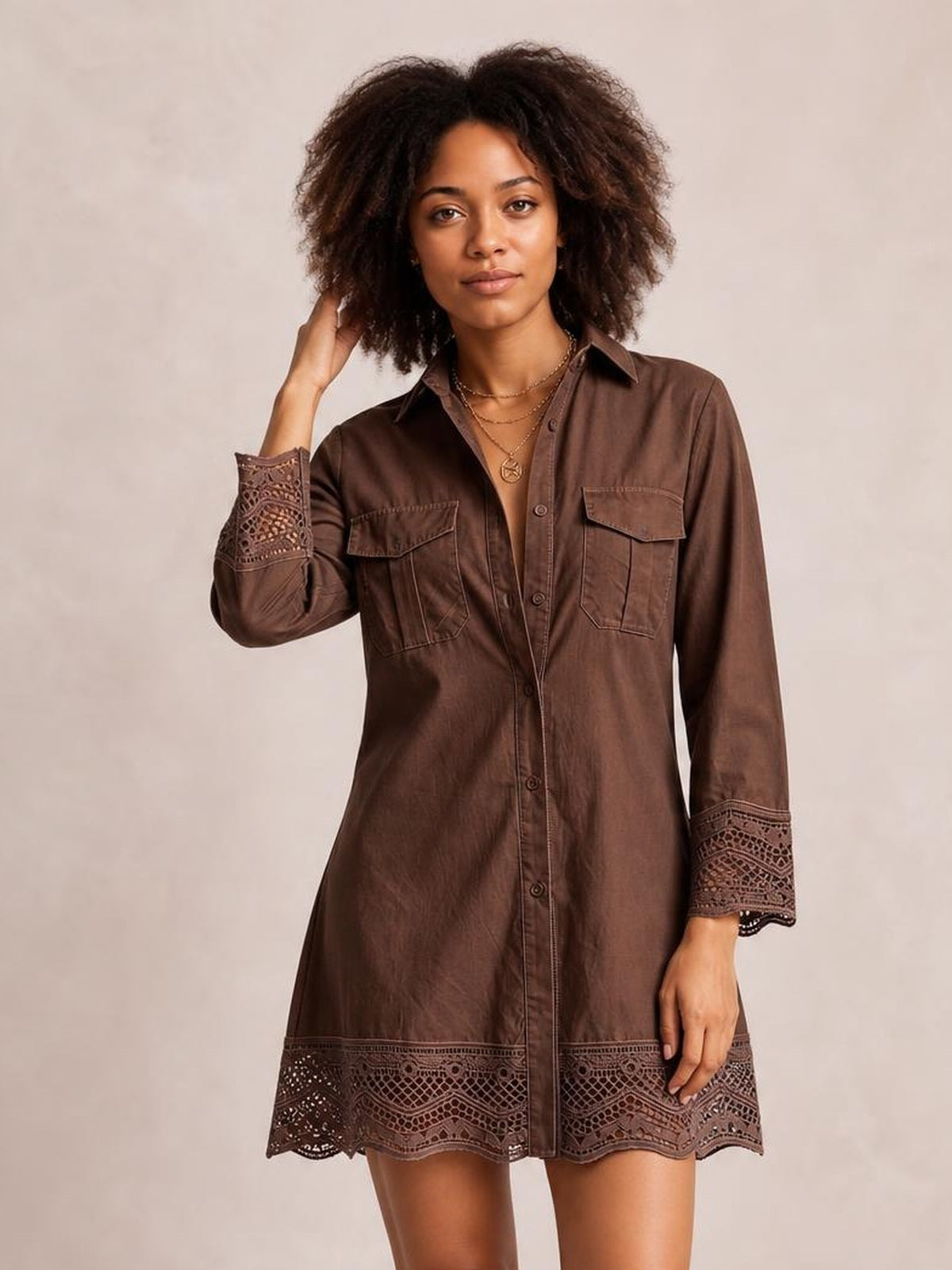 Model poses with one hand in her hair wearing a brown button-down shirt dress with chest pockets, a relaxed silhouette, and lace-trimmed cuffs and hem.