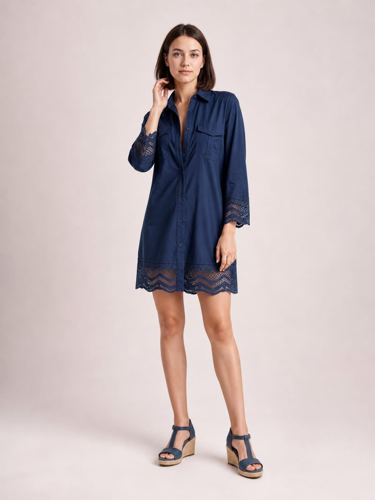Model wearing a navy button-down shirt dress with chest pockets, lace-trimmed eyelet cuffs and hem, a straight short silhouette, and one hand touching her neck.