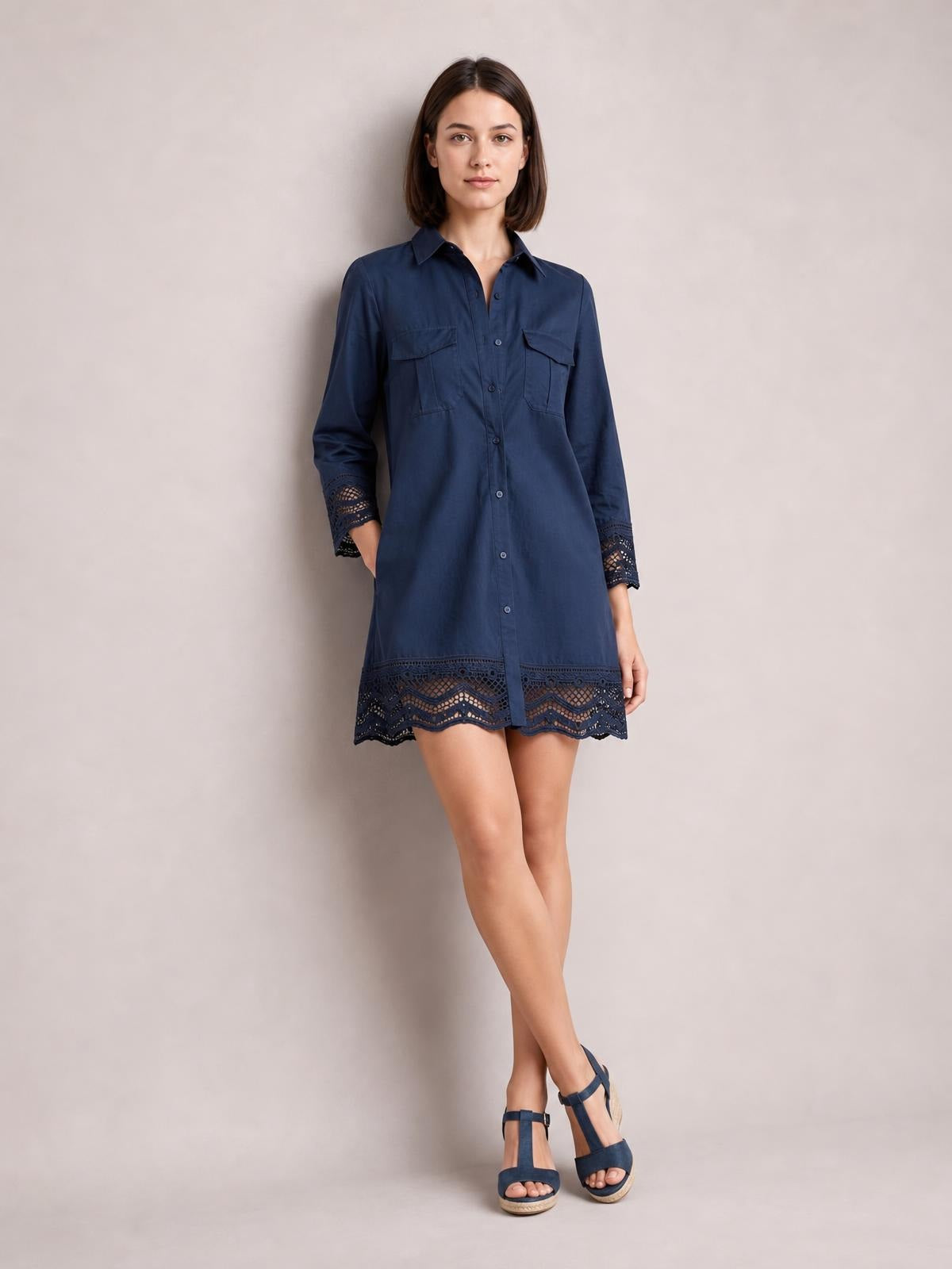 Model poses in a navy button-down shirt dress with a relaxed silhouette, chest pockets, long sleeves, and eyelet lace trim at the cuffs and hem.