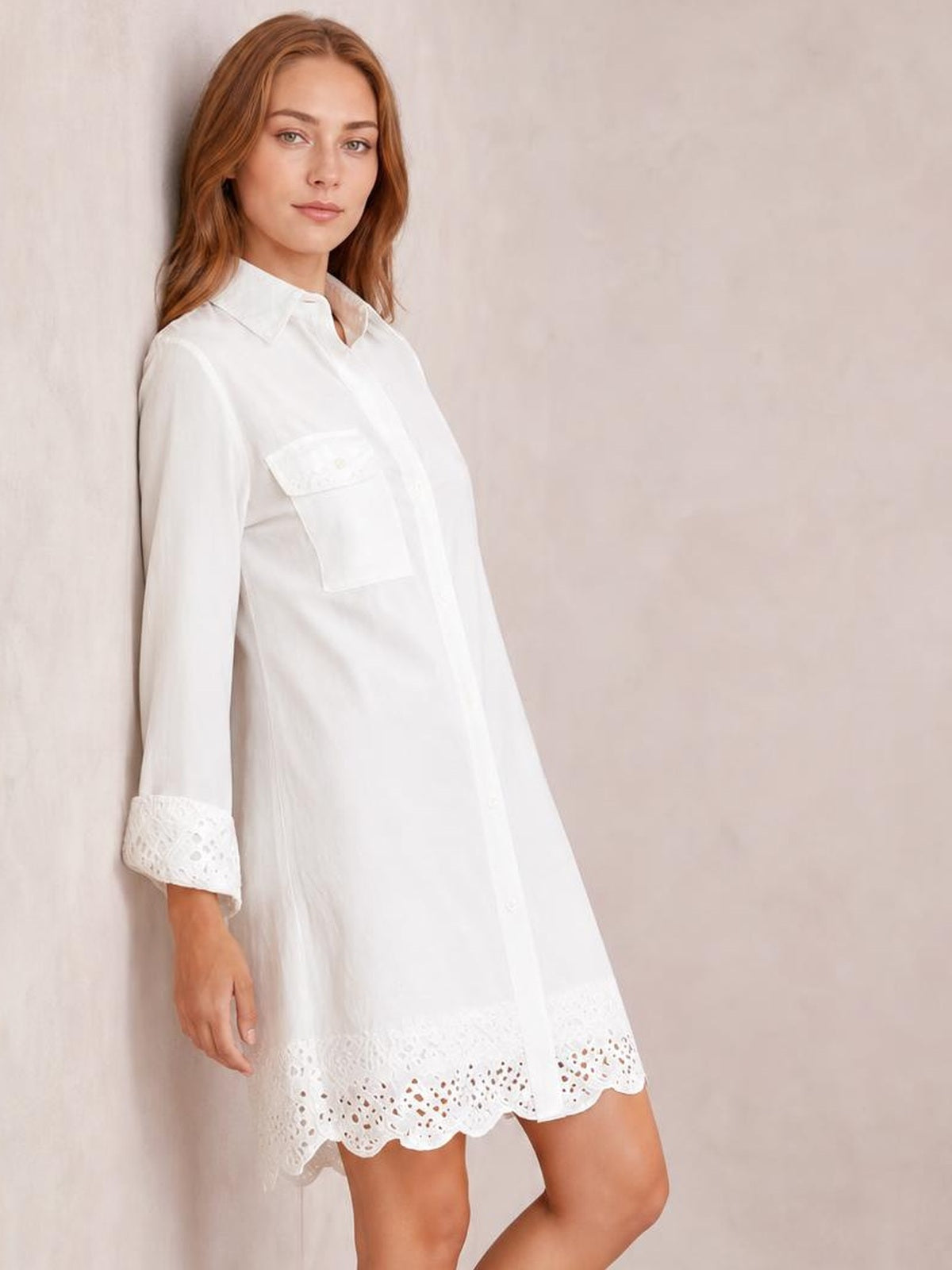 Model leans against a wall in a white button-down shirt dress with a collared neckline, chest pocket, long sleeves, lace-trimmed cuffs, and scalloped eyelet hem.