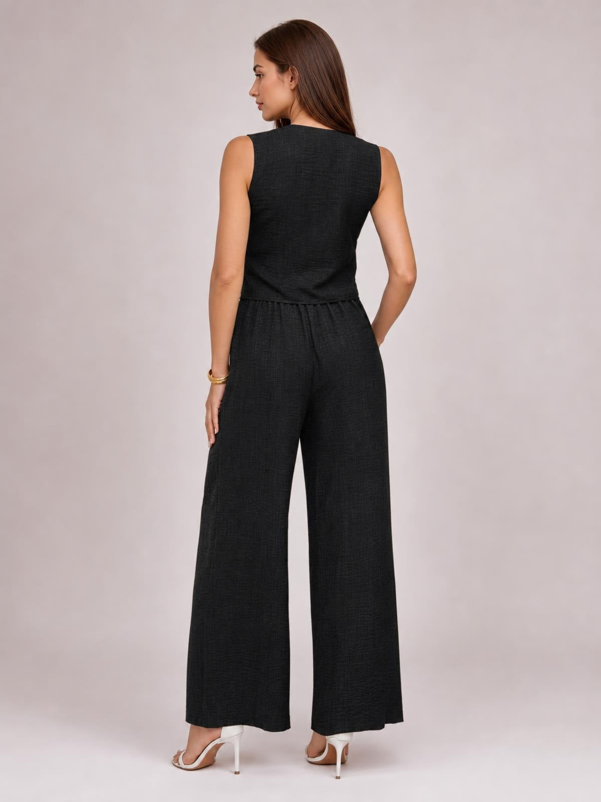 Model poses in a black sleeveless button-down vest and matching wide-leg pants set, shown from the back with a relaxed, tailored silhouette and pockets.