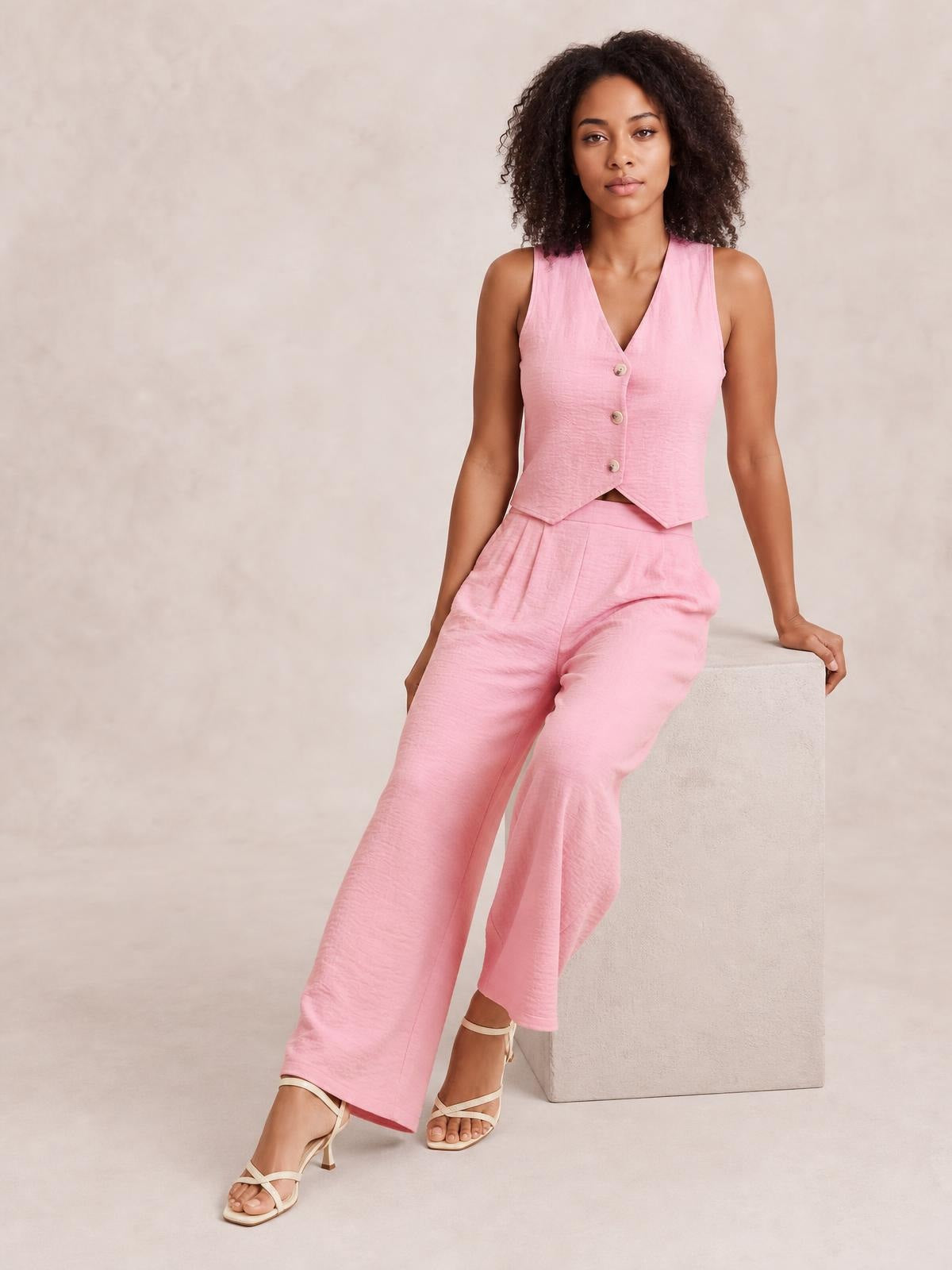 A model poses seated in a pink button-down vest and matching wide-leg pants set, featuring a tailored sleeveless silhouette and front buttons.
