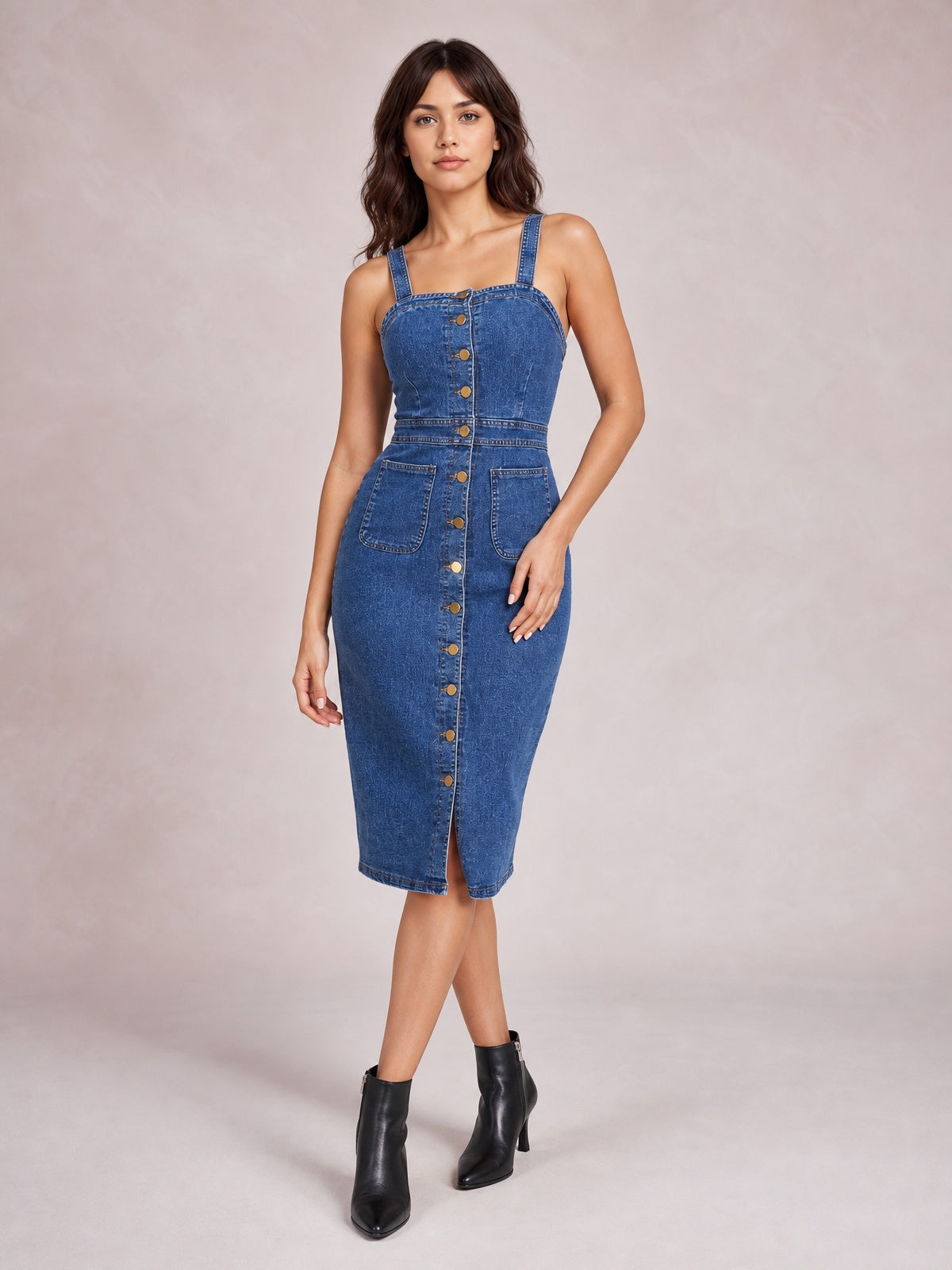 A model poses in a dark blue fitted denim midi dress with wide straps, a softly square neckline, defined seams, button front, and two patch pockets.