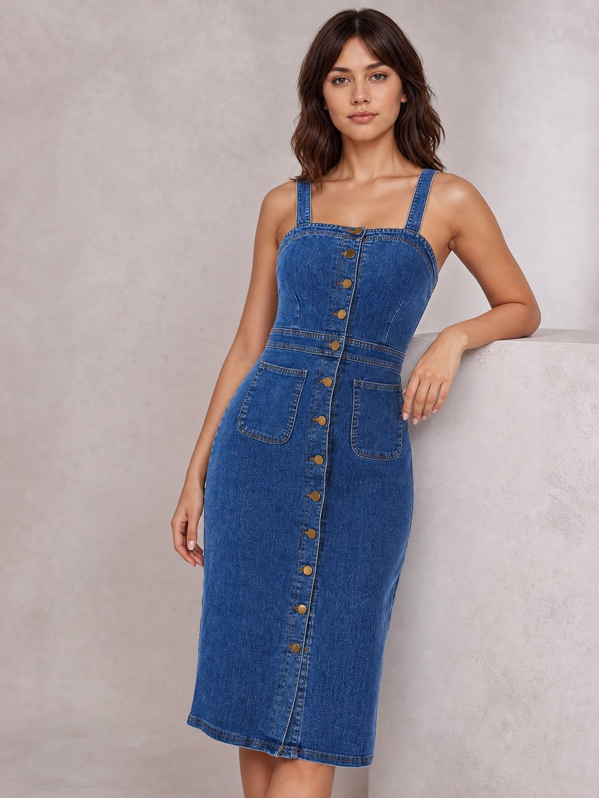 Model wearing a dark blue fitted denim midi dress with wide straps, a squared neckline, defined seams, button-front closure, and two patch pockets.