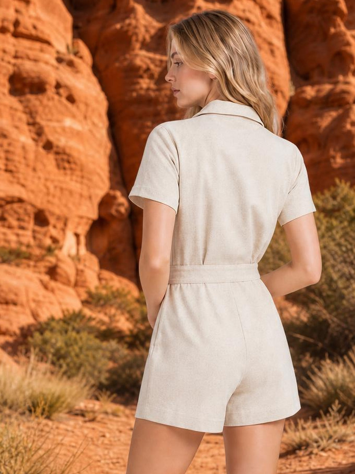 A model stands turned away in a cream button-front romper with short sleeves, a pointed collar, defined waist seam, and relaxed wide-leg shorts.