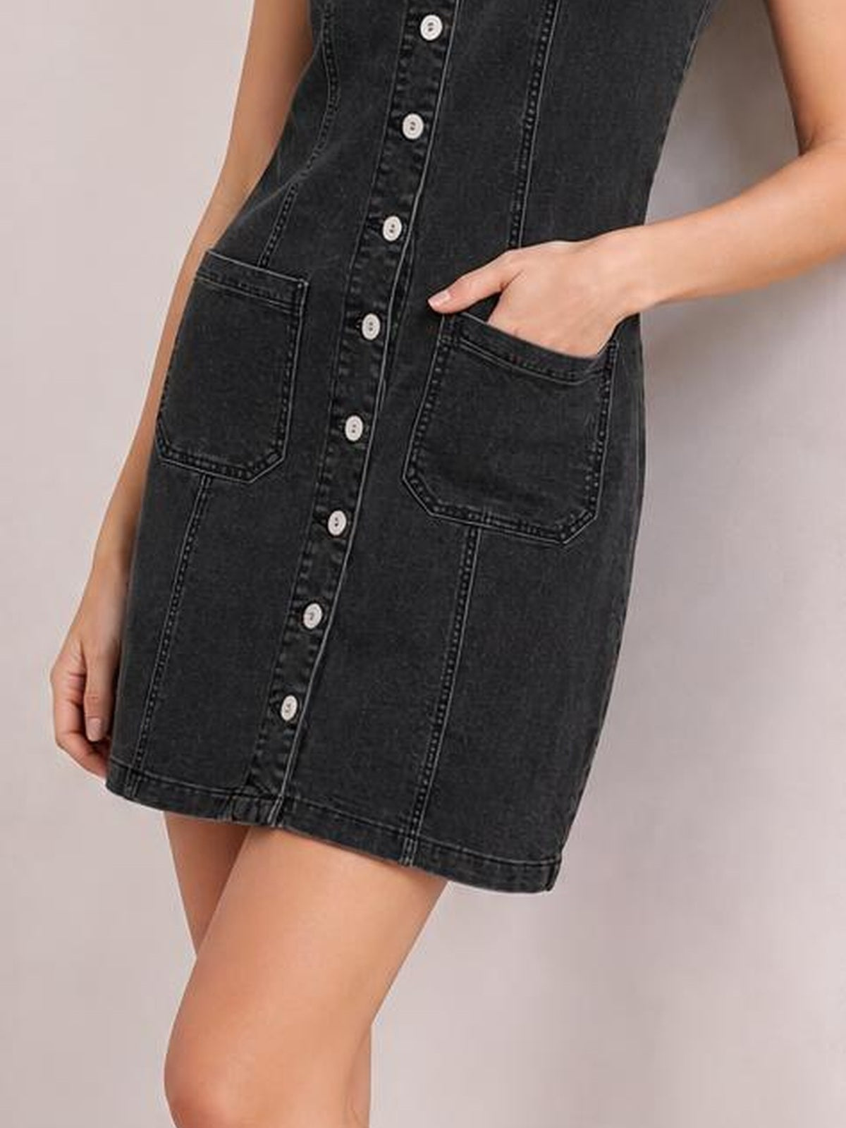 Model in a sleeveless black denim dress with a button-front, straight mini silhouette, contrast stitching, and two large patch pockets, one hand tucked inside.