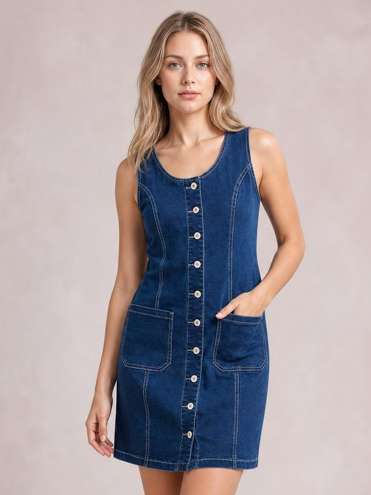 Model poses in a dark blue sleeveless denim dress with a round neckline, button-front closure, contrast stitching, fitted silhouette, and two patch pockets.