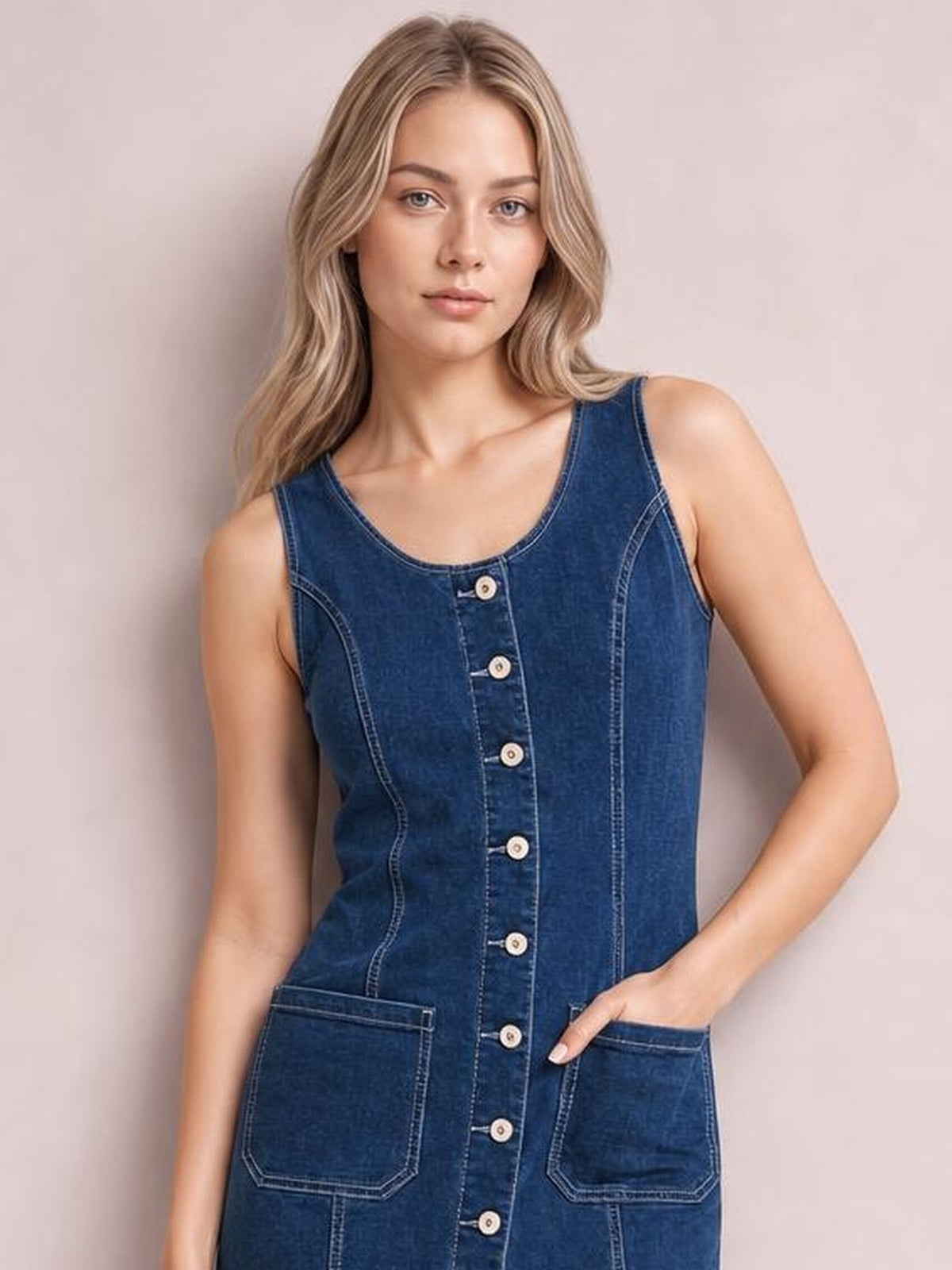 Model poses in a dark blue sleeveless denim dress with a scoop neckline, button-front closure, curved seams, and two large patch pockets.