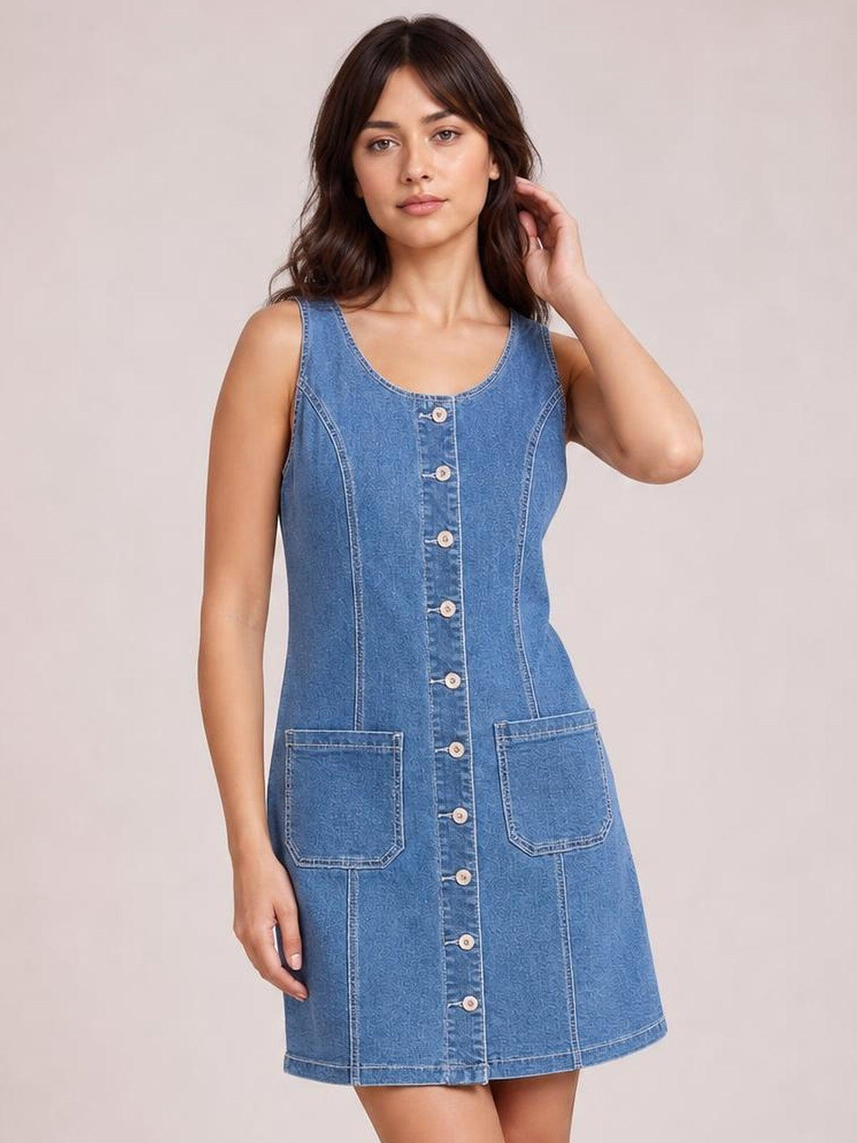 Model poses in a blue sleeveless denim dress with a scoop neckline, button-front closure, fitted waist, contrast seams, two patch pockets, and an A-line silhouette.