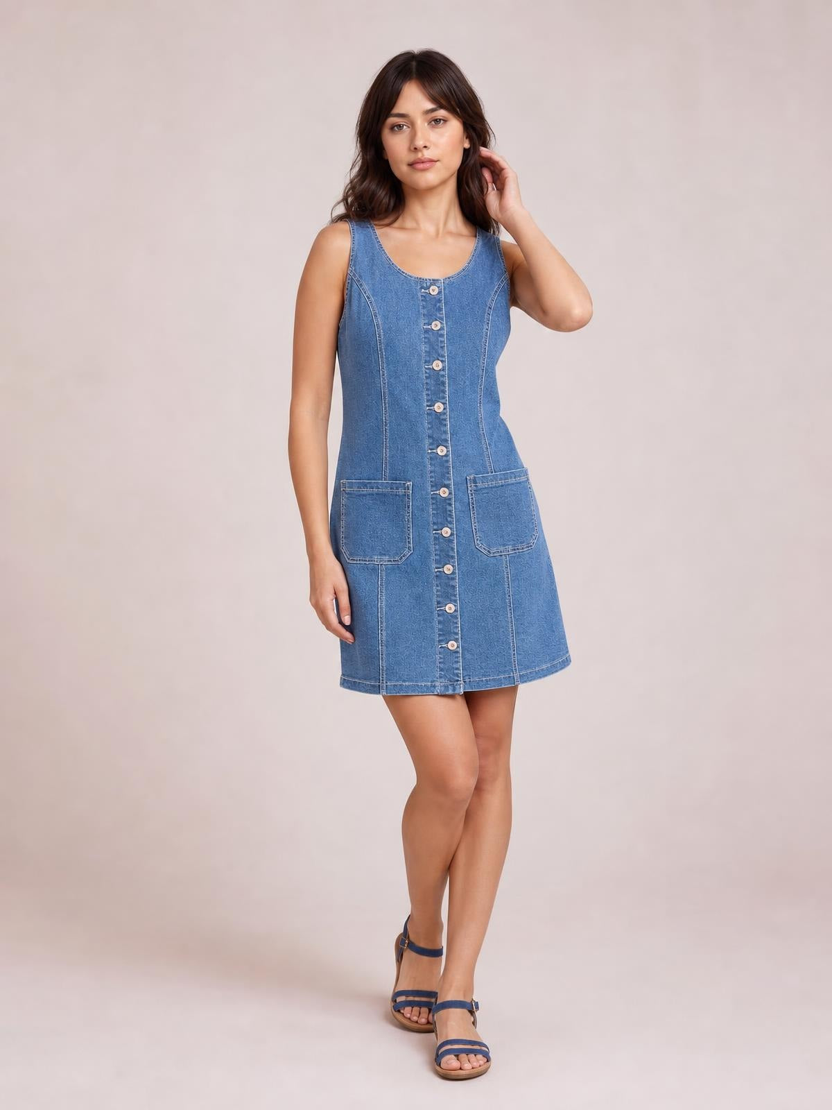 Model poses in a blue sleeveless denim dress with a scoop neckline, button-front construction, curved seams, two patch pockets, and a fitted A-line silhouette.