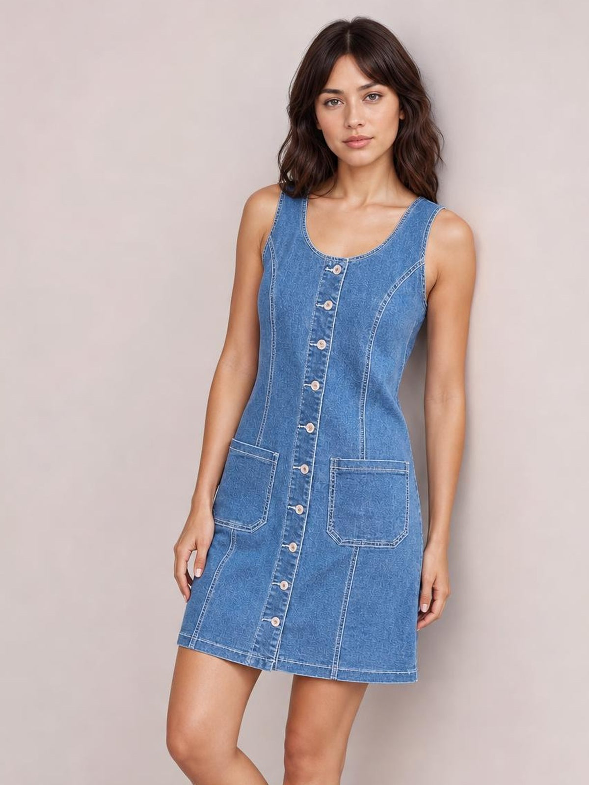 Model wearing a blue sleeveless denim dress with a scoop neckline, button-front closure, fitted seamed silhouette, contrast stitching, and two large front pockets.