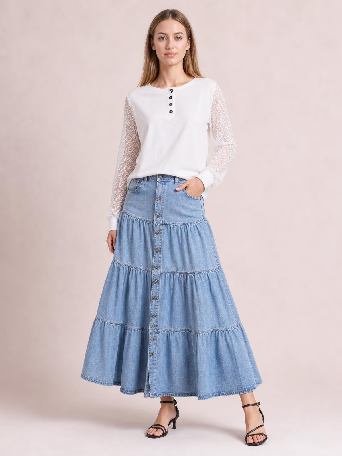 Model wears a light blue denim skirt with a button-front placket, multiple gathered tiers, side pockets, and a full flowing silhouette, posing with one hand tucked in a pocket.