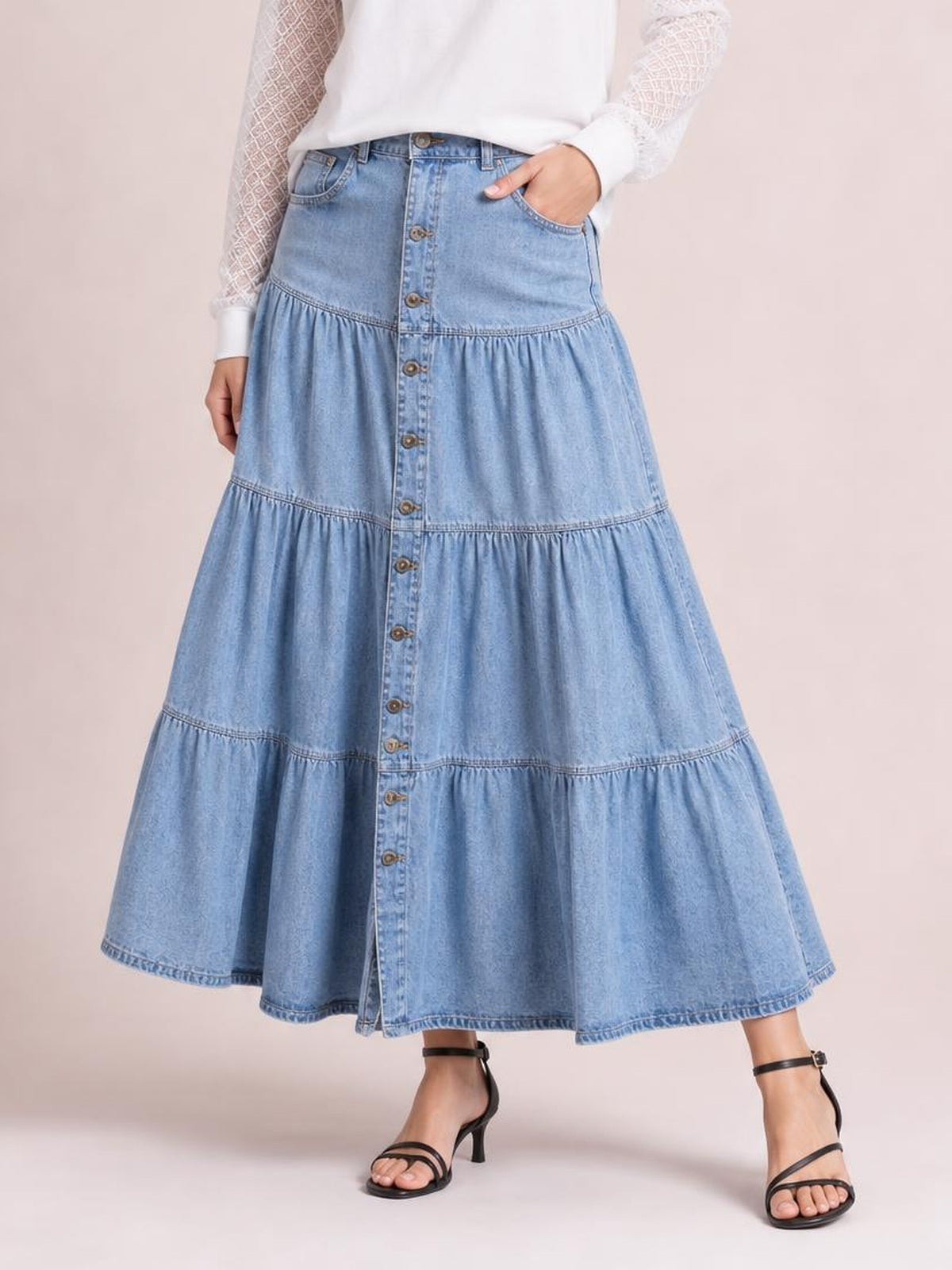 A model wears a light blue denim skirt with a button front, multiple gathered tiers, side pockets, and a flowing A-line silhouette.
