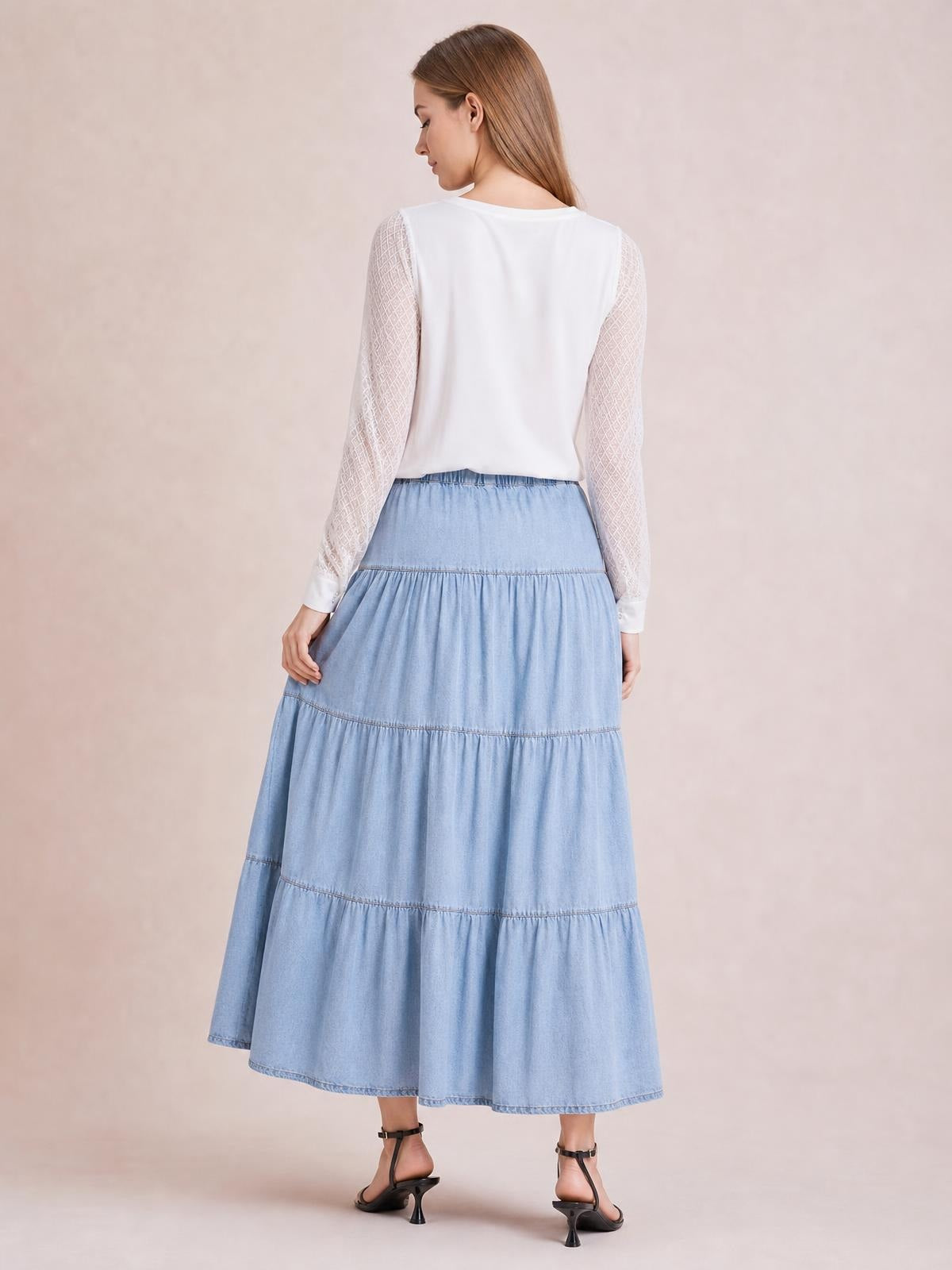 Model shown from behind in a light blue tiered denim skirt with a gently gathered waistband, multiple seamed panels, and a flowing midi silhouette.