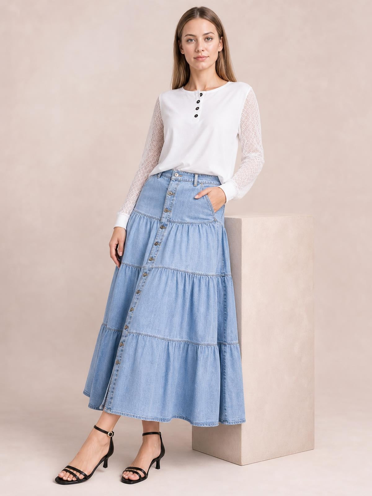 Model poses in a light blue denim button-front tiered midi skirt with visible pockets and a relaxed, flowing silhouette.