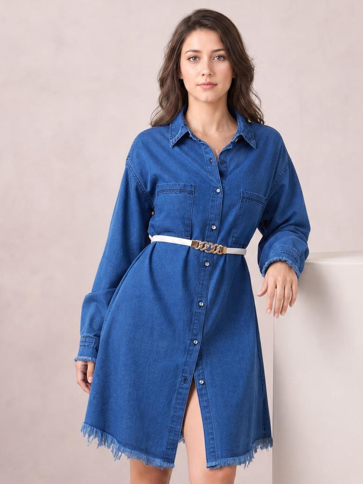 Model poses in a blue button-up collared neck denim dress with long sleeves, chest pockets, a relaxed silhouette, and a raw fringed hem.