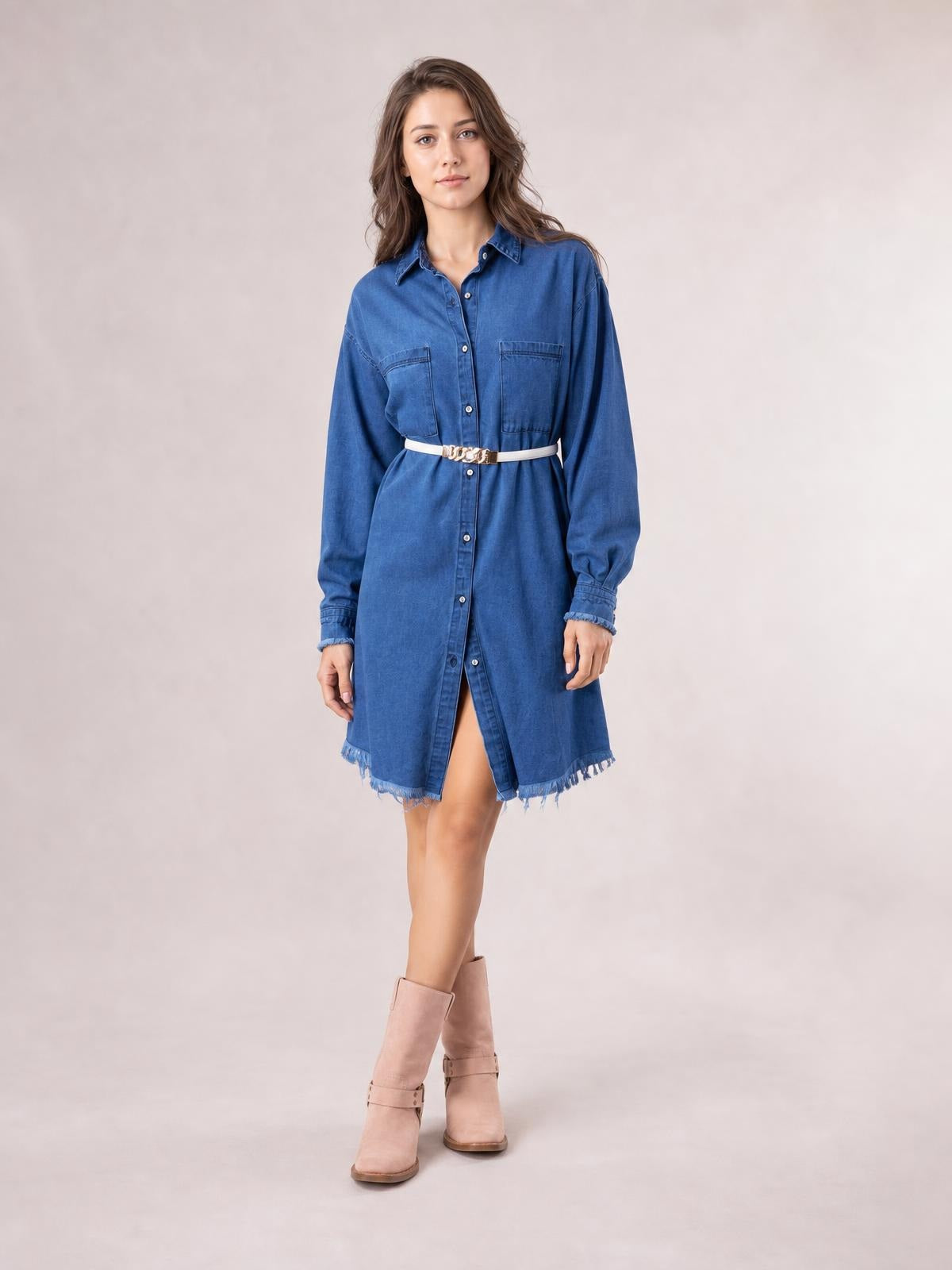 A model poses in a blue button-up collared denim dress with long sleeves, chest pockets, a relaxed straight silhouette, and a raw frayed hem.