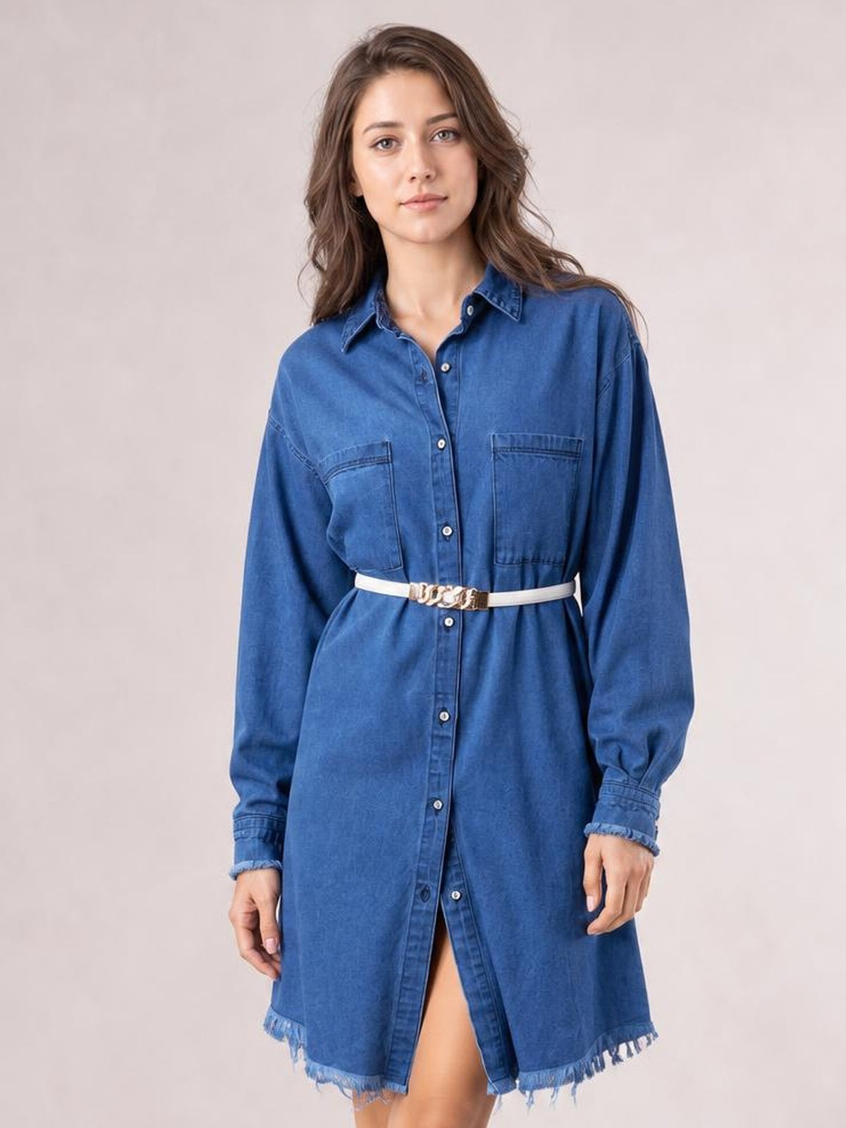 A model stands in a blue denim dress with a collared neckline, button-front construction, long sleeves, patch pockets, relaxed silhouette, and raw frayed hem.