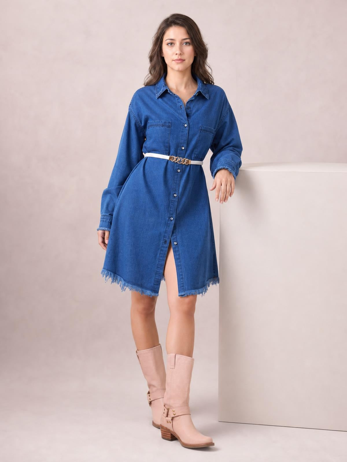 A model stands in a blue button-up collared neck denim dress with long sleeves, chest pockets, a relaxed silhouette, waist belt, and raw frayed hem.