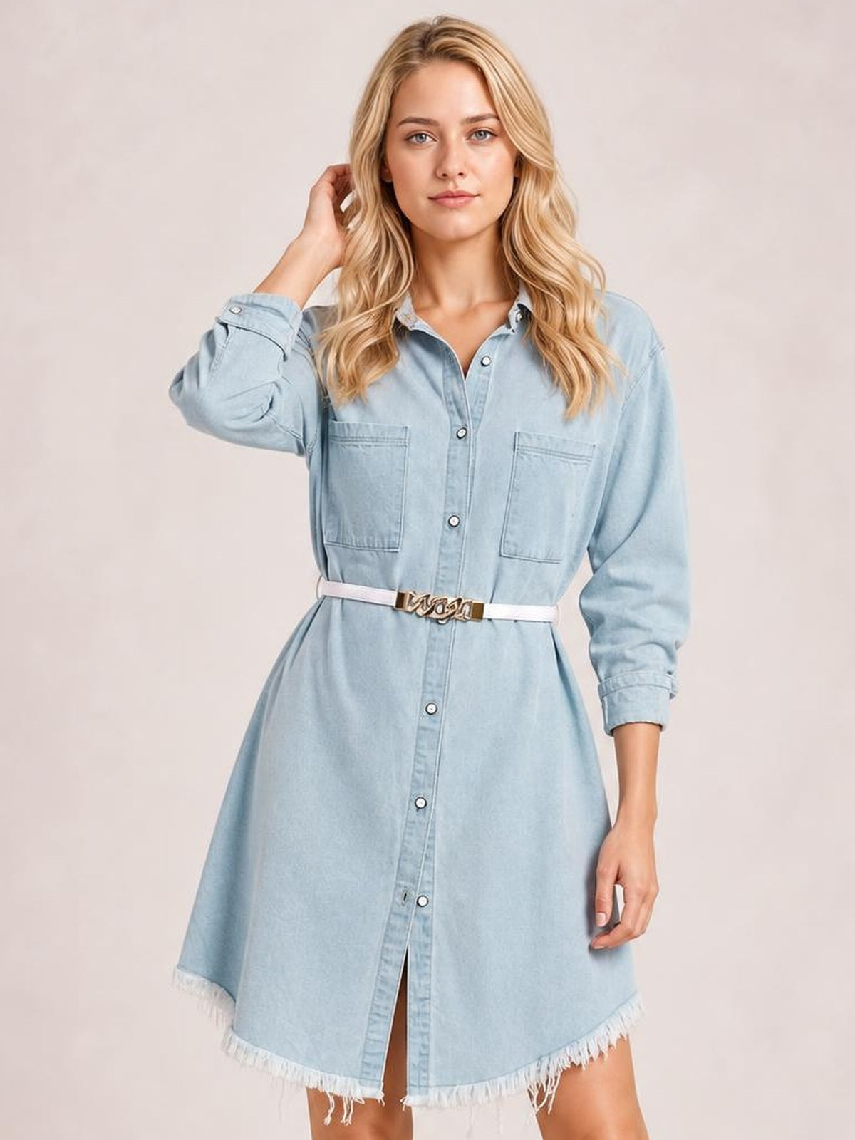 A model poses in a light blue denim dress with a collared button-up front, chest pockets, long sleeves, belted waist, and raw hem.
