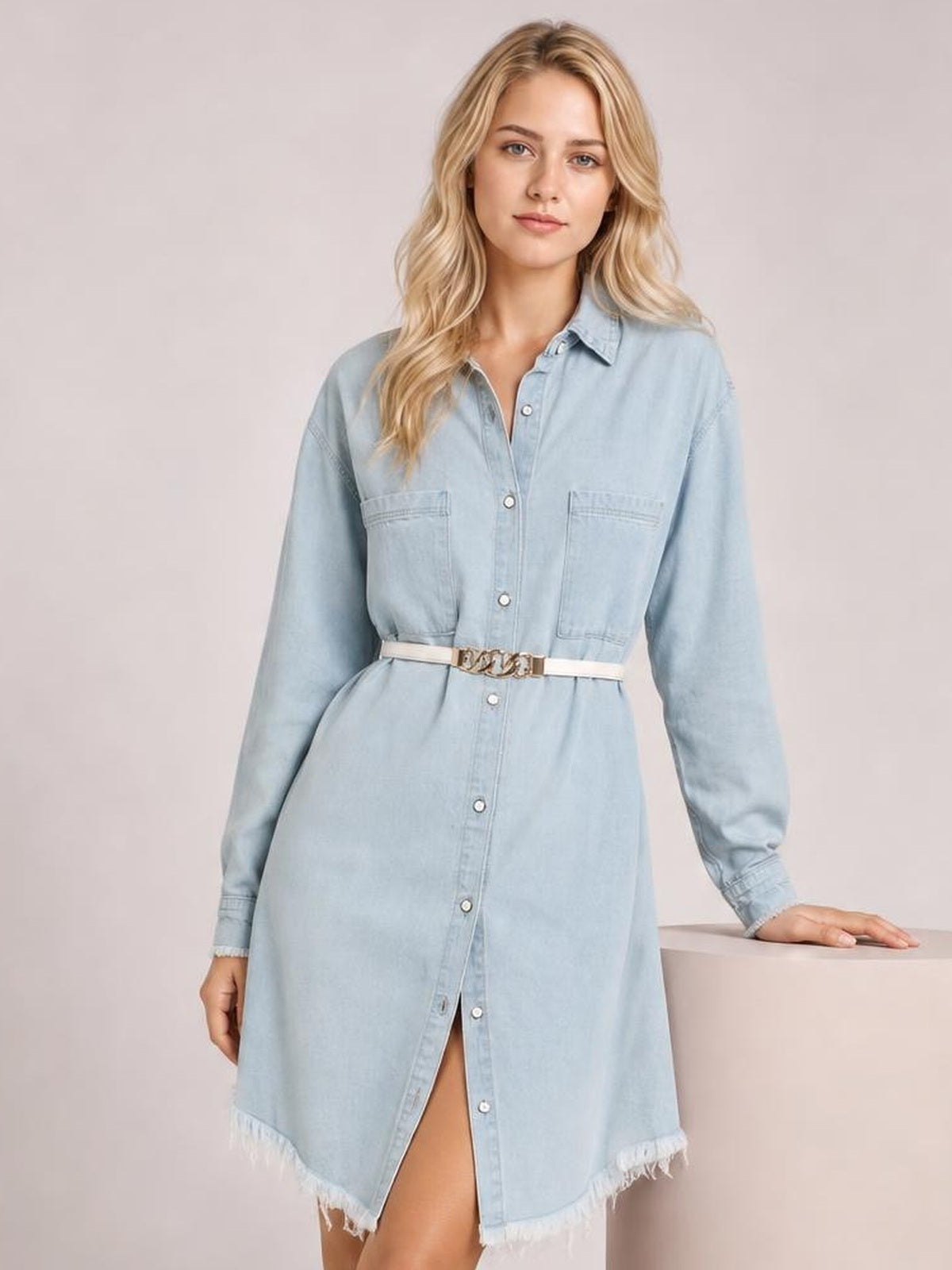 Model wears a light blue button-up collared neck denim dress with long sleeves, chest pockets, a belted waist, straight silhouette, and raw hem.