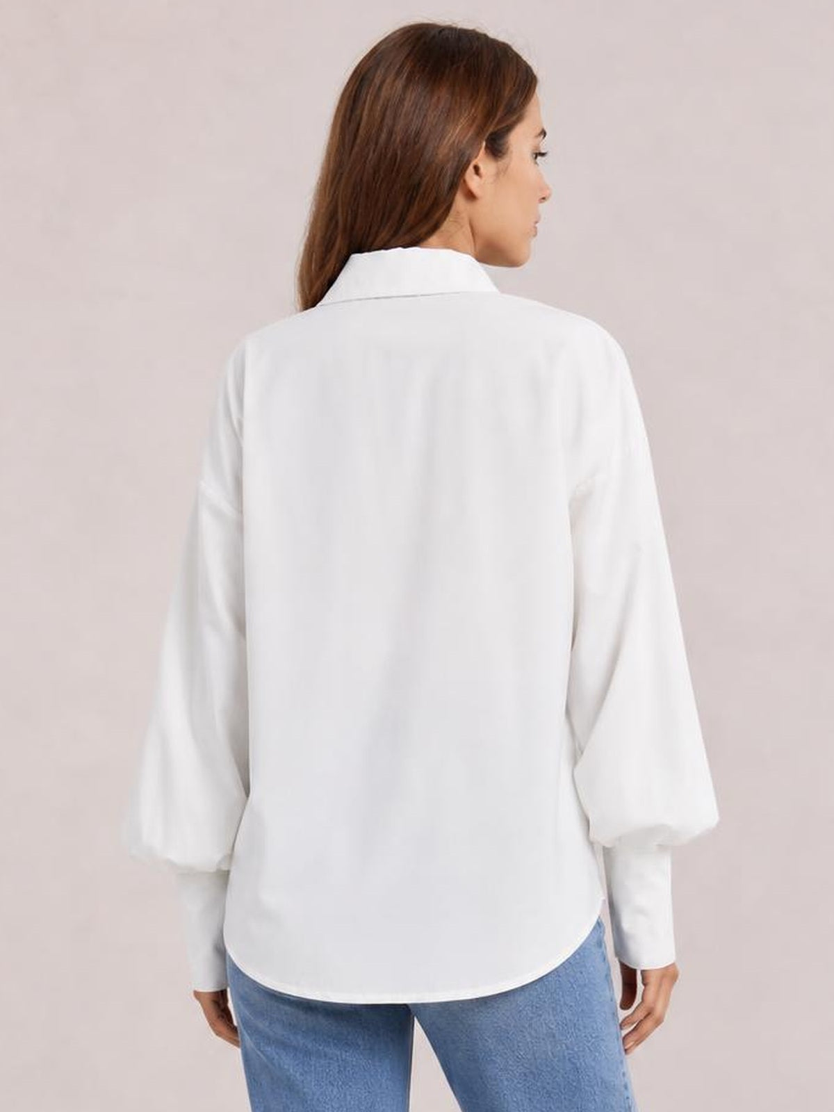 A model turns slightly to show a white button-up shirt with a collared back, dropped shoulders, loose silhouette, and voluminous lantern sleeves.