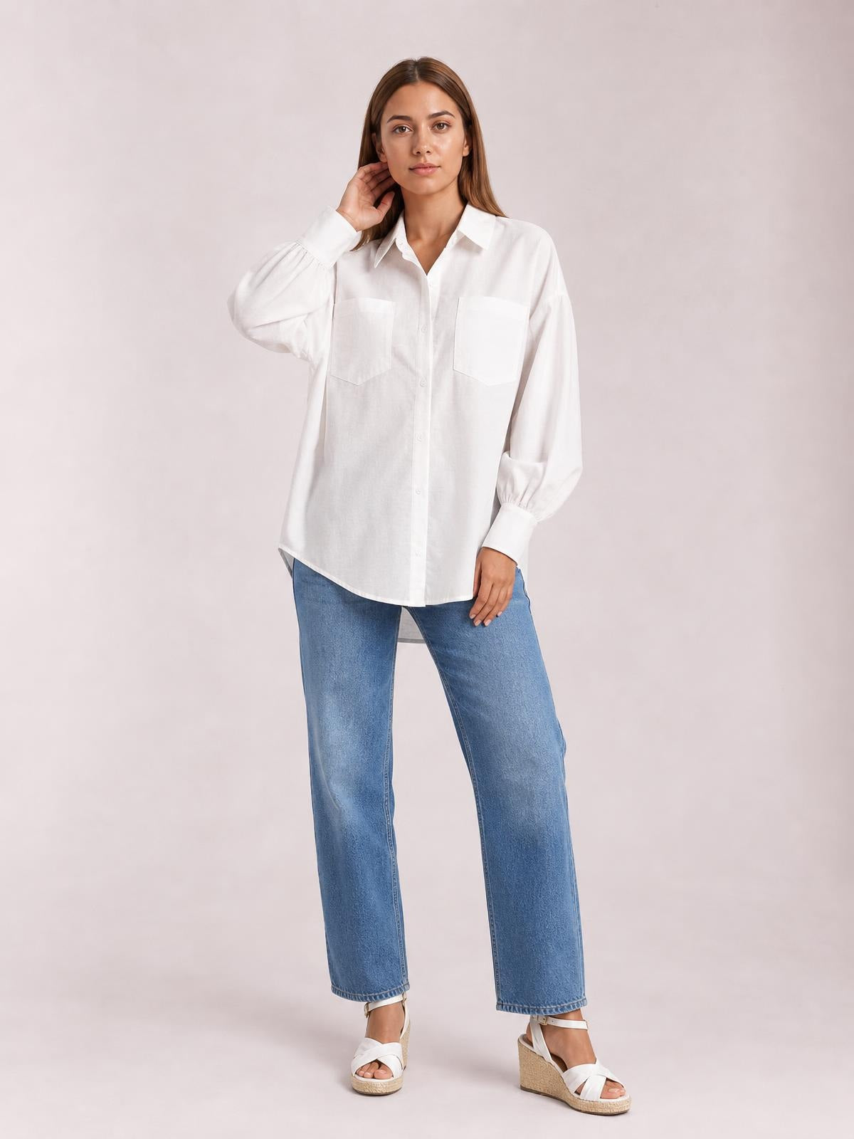 A woman poses in a white button-up shirt with dropped shoulders, voluminous lantern sleeves, two chest pockets, and a relaxed longline silhouette.