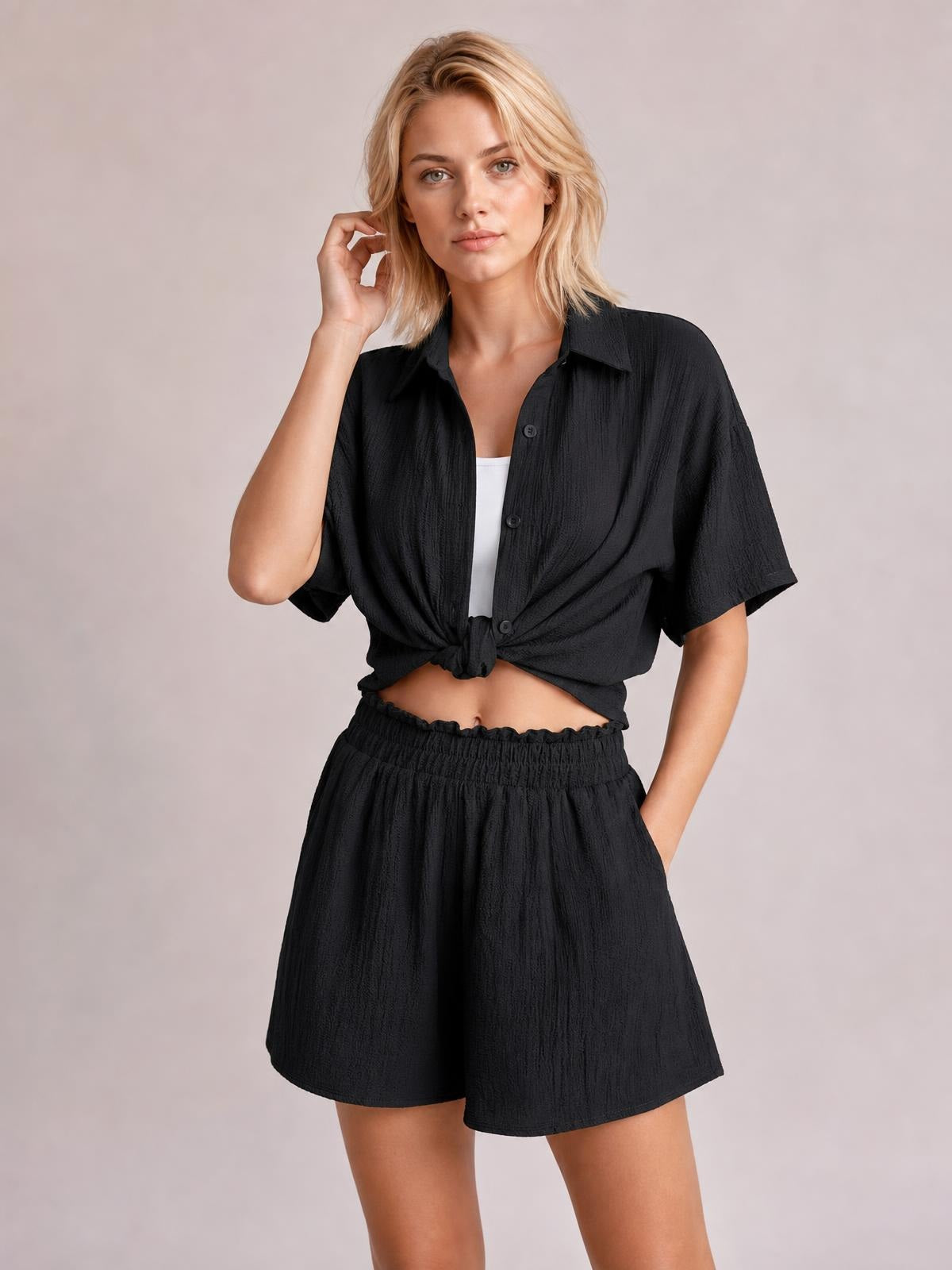 Model poses in a black button-up half-sleeve top tied at the waist with matching pocketed shorts, an elastic waistband, and a relaxed fit.