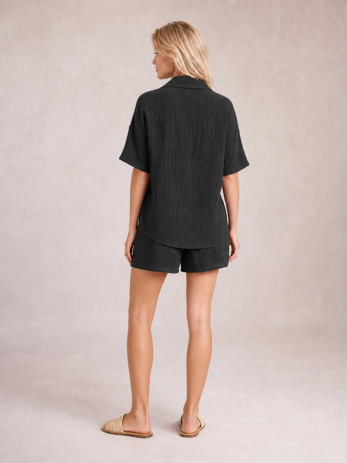 Model shown from behind wearing a black button-up half-sleeve top and matching shorts set with a relaxed silhouette and curved shirt hem.