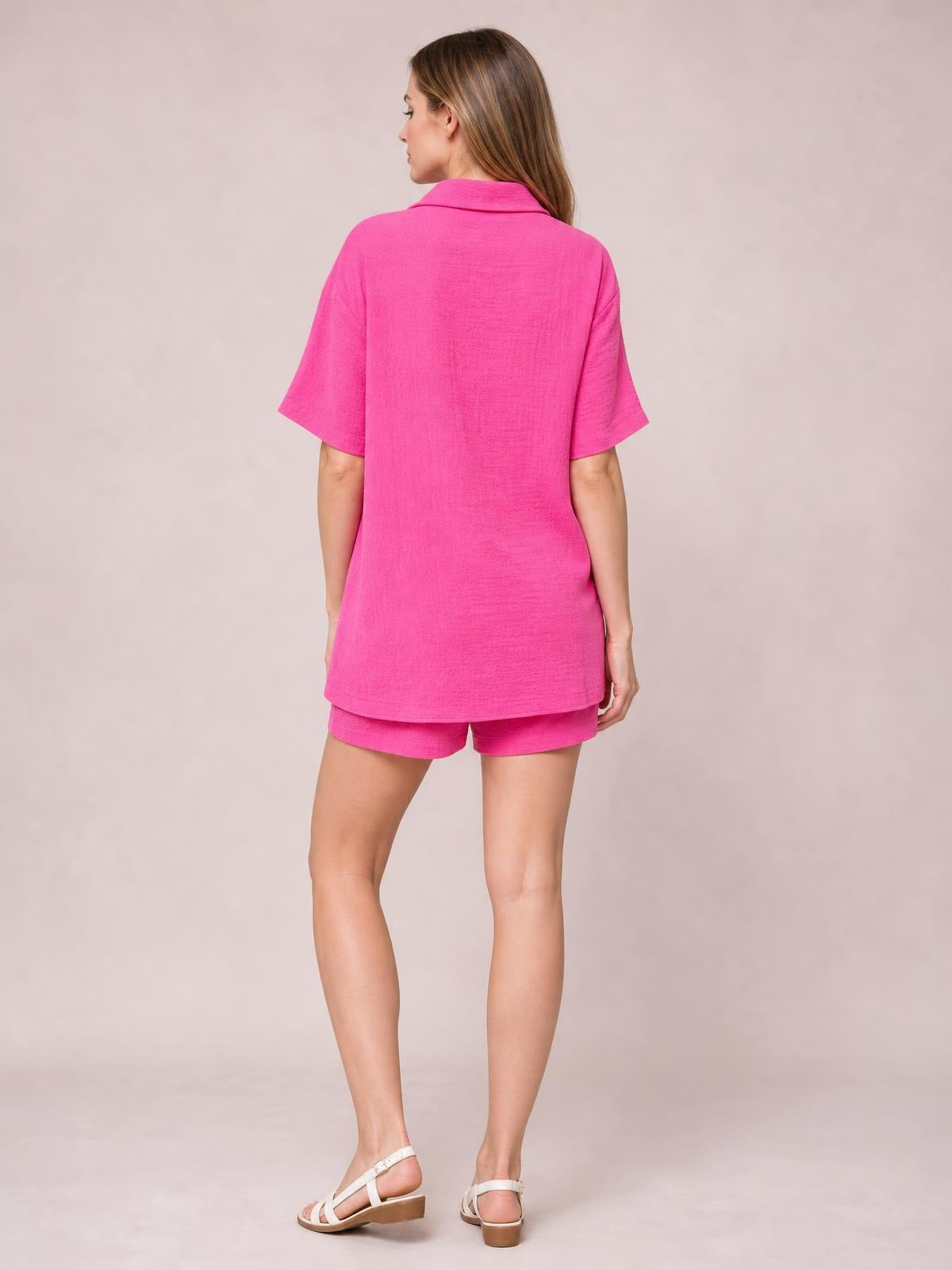 Model turned away wears a bright pink button-up half-sleeve top with a relaxed longline silhouette and matching shorts in this two-piece set.
