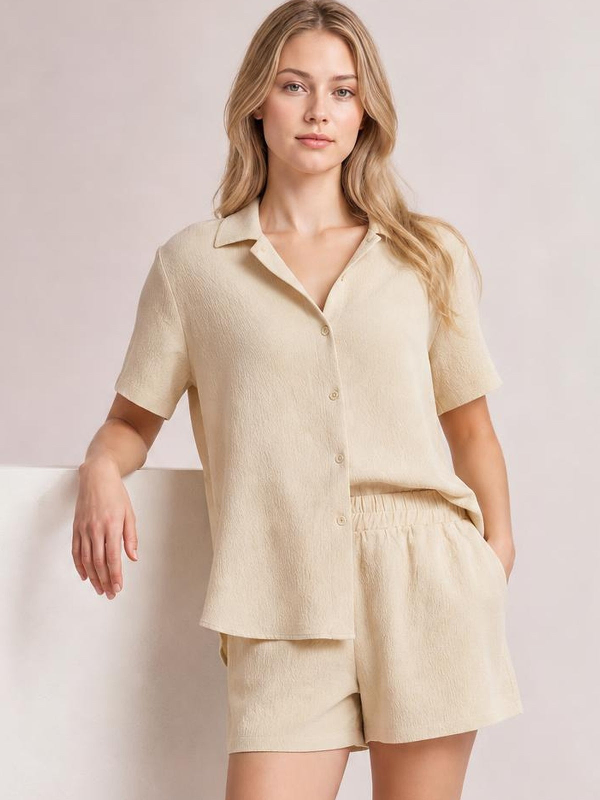 Model poses in a cream button-up half-sleeve top and matching shorts set, featuring a collared relaxed silhouette, elastic waistband, and side pockets.