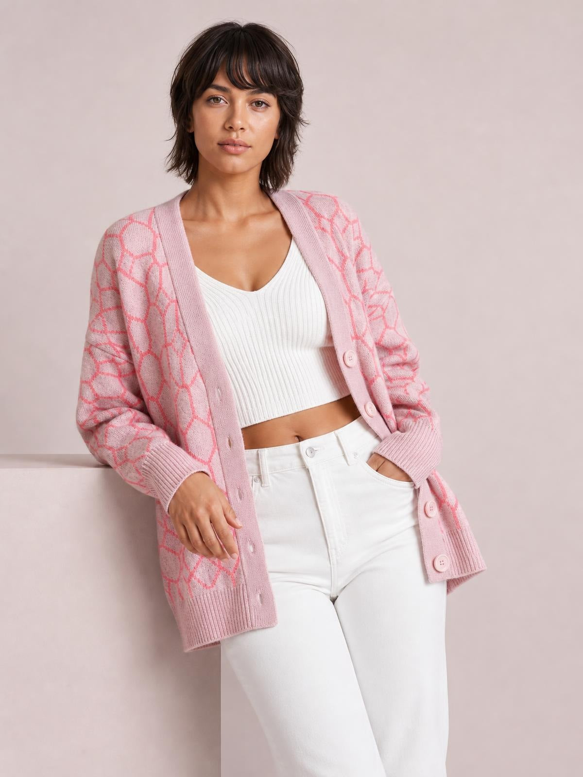 Model wears a pink long sleeve cardigan with a button-up front, geometric pattern, ribbed trim, and relaxed longline silhouette over a white top.