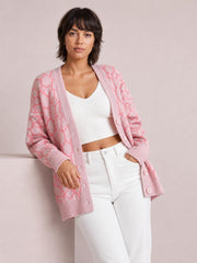 Model wears a pink long sleeve cardigan with a button-up front, geometric pattern, ribbed trim, and relaxed longline silhouette over a white top.