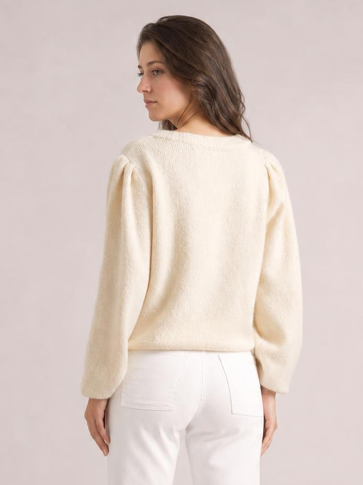 Model looks over her shoulder wearing a cream knit cardigan with long sleeves, a rounded neckline, softly puffed shoulders, and a relaxed straight silhouette.