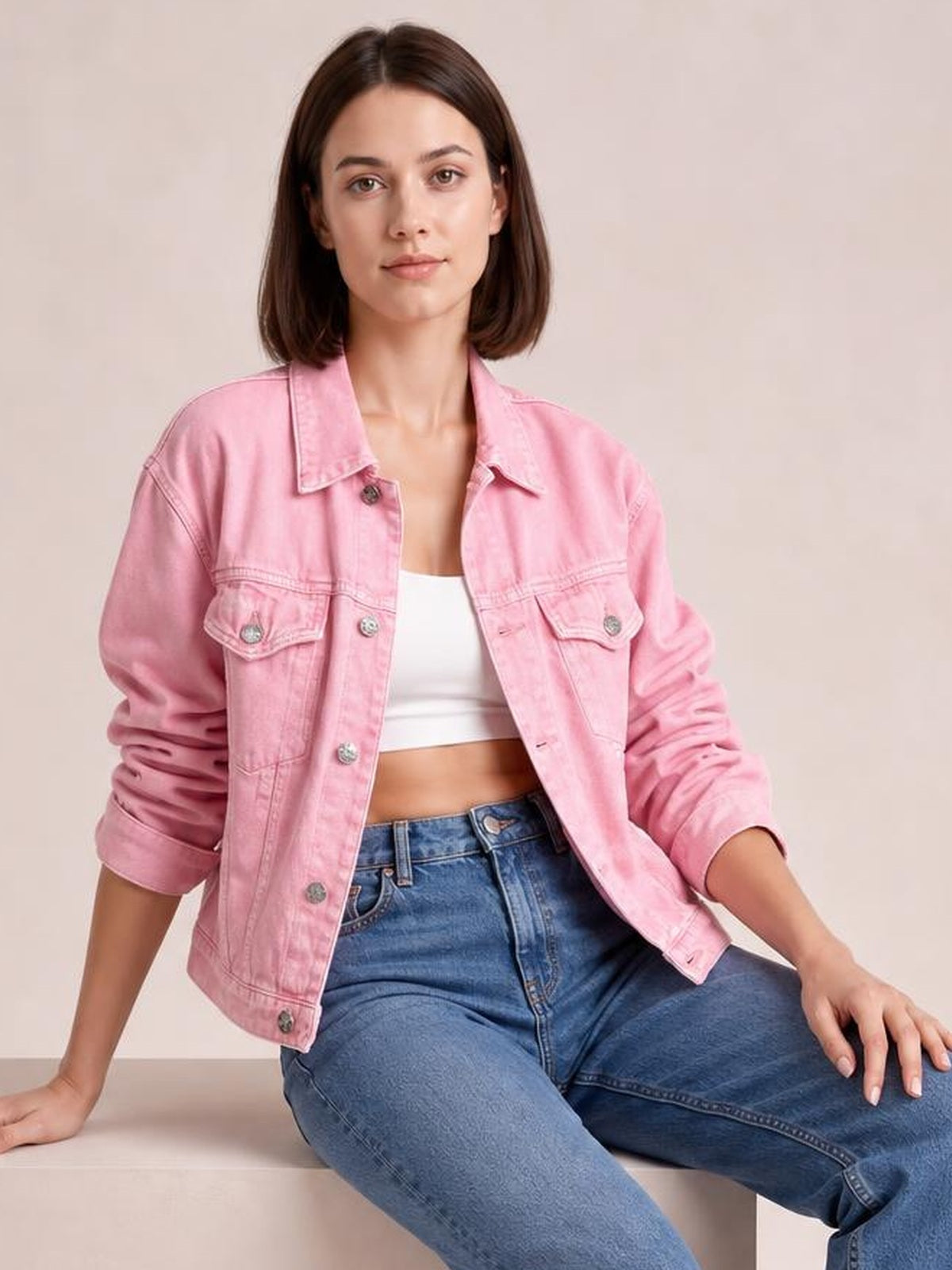 A seated model wears a pink button-up long sleeve denim jacket with a collared front, chest flap pockets, and a relaxed silhouette.