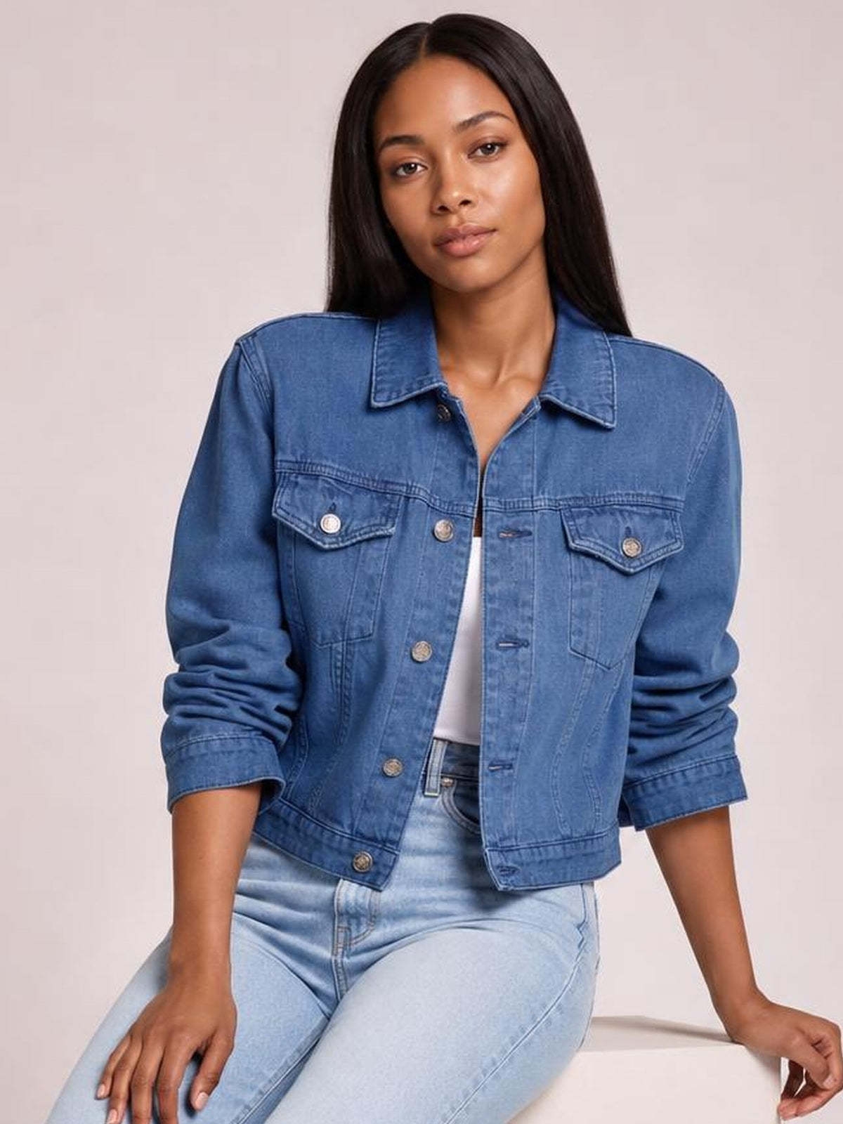 Model seated in a blue button-up long sleeve denim jacket with a structured, waist-length silhouette, pointed collar, and flap chest pockets.