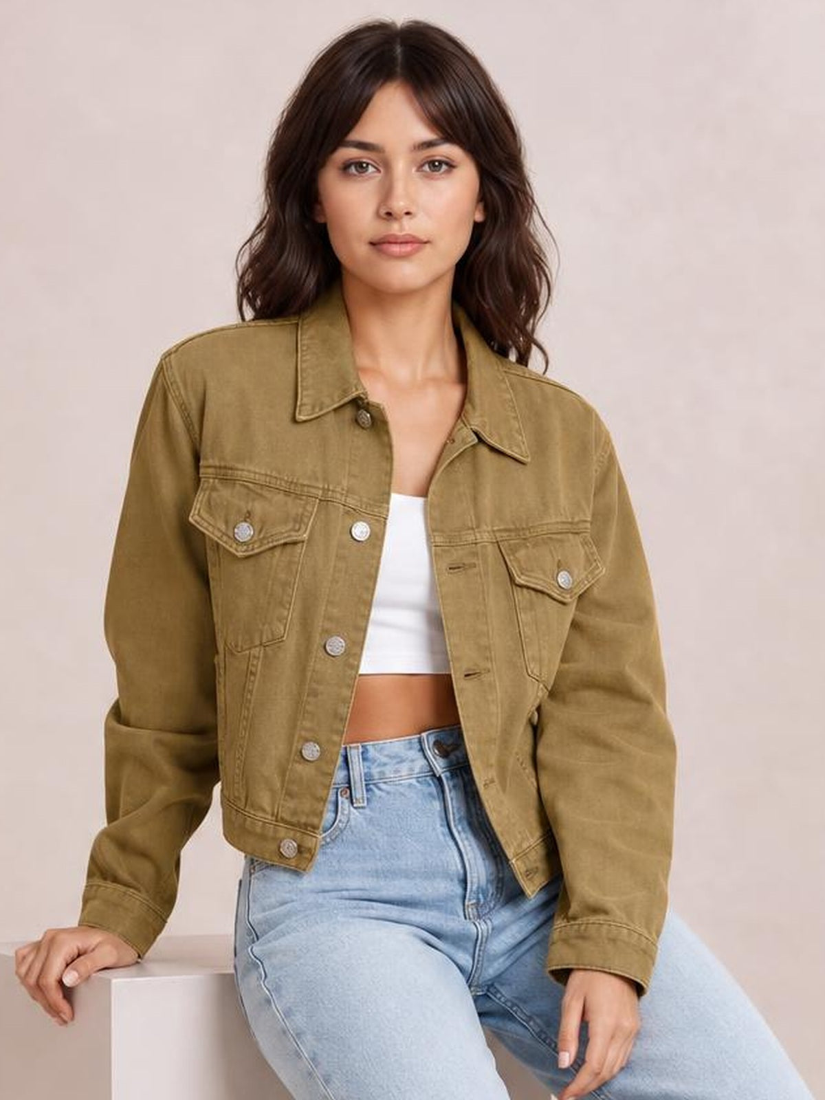 Model seated in a tan button-up long sleeve denim jacket with a collared neckline, cropped silhouette, chest flap pockets, and visible metal buttons.
