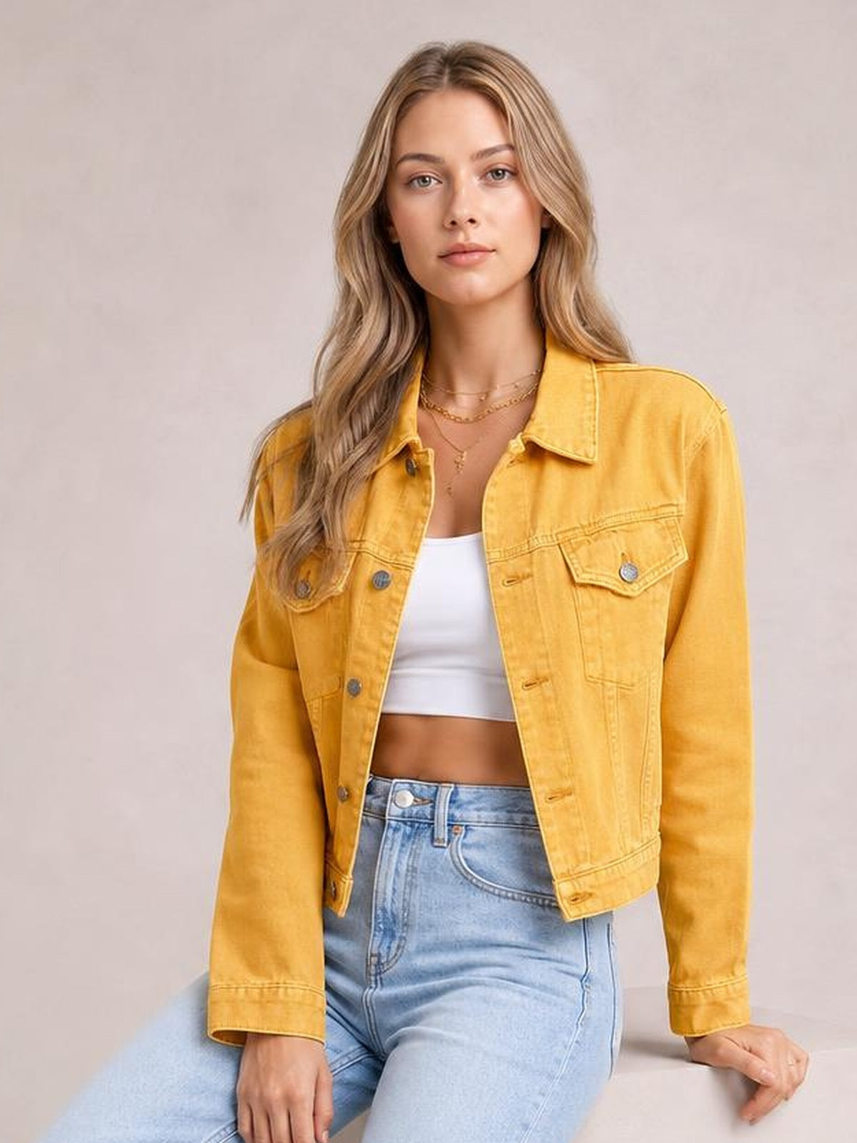 A model sits in a mustard-yellow button-up long sleeve denim jacket with a relaxed waist-length silhouette and two flap chest pockets.